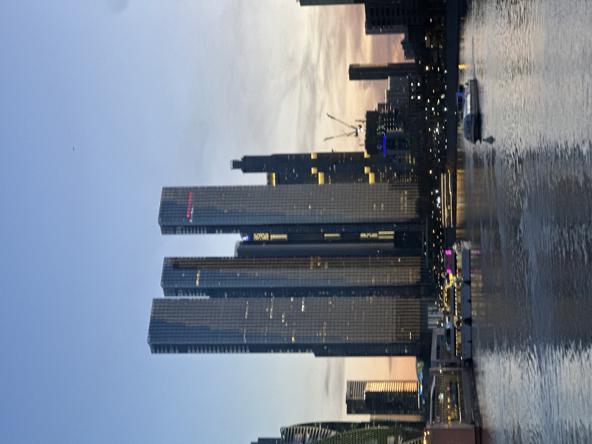 The image captures a vibrant city skyline at dusk or early evening, showing a cluster of modern skyscrapers alongside a wide body of water, likely a river. The scene is illuminated by the fading light of the sky and the artificial lights of the buildings.
In the foreground, a river with a rippled surface reflects the soft, warm hues of the sky. A white and dark-colored boat is visible on the water, creating a distinct wake behind it, indicating movement.
Dominating the midground are several tall, glass-fronted skyscrapers. Many windows are illuminated, casting a warm glow from within the dark facades. On one prominent building, red text, possibly Cyrillic, is visible near its top edge, suggesting a corporate name like "СБЕР" (Sberbank). Another building displays a yellow "24" sign. Further in the background, more city buildings extend into the distance, with construction cranes visible against the sky, suggesting ongoing development.
The sky transitions from a clear, light blue at the top to a warm, hazy band of orange and pink near the horizon, indicative of twilight. A small, dark speck, possibly a bird or drone, is visible high in the sky. The overall impression is one of a bustling urban environment transitioning from day to night.