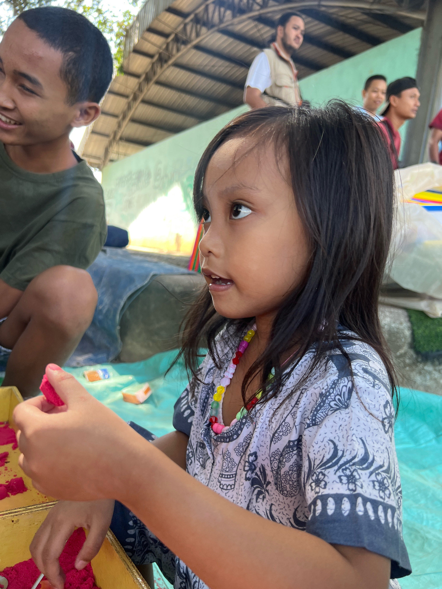 A young girl with dark hair and a multicolored beaded necklace plays with pink kinetic sand in a golden box, her left hand holding a small pink piece. She wears a patterned shirt. To her left, a young boy with short dark hair smiles, wearing a green t-shirt. In the background, several other young men and an adult male in a vest are visible under a corrugated metal roof. The scene appears to be outdoors, possibly in a public space like a community center or school in Kecamatan Pati, Indonesia, with a light blue tarp covering the ground.