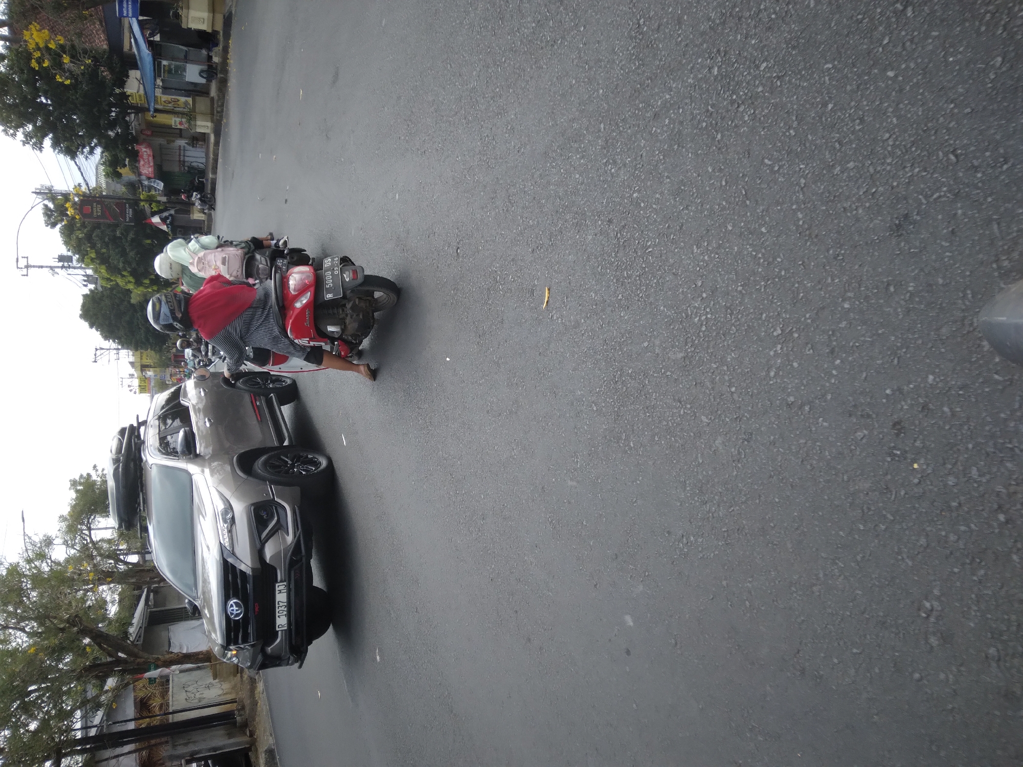 The image depicts a street scene in Purwokerto, Indonesia. A silver Toyota Fortuner SUV with a roof box is driving on the left side of the road, displaying a license plate "R 1937 MJ". In front of the SUV, two individuals are riding a red scooter. The rider in front, wearing a red hijab and a striped shirt, is looking back. The passenger behind her is wearing a white hijab and a light pink backpack. The scooter has a license plate "R 5000 DS". The street is asphalt, and there are trees and buildings lining the right side of the road. The sky is overcast.