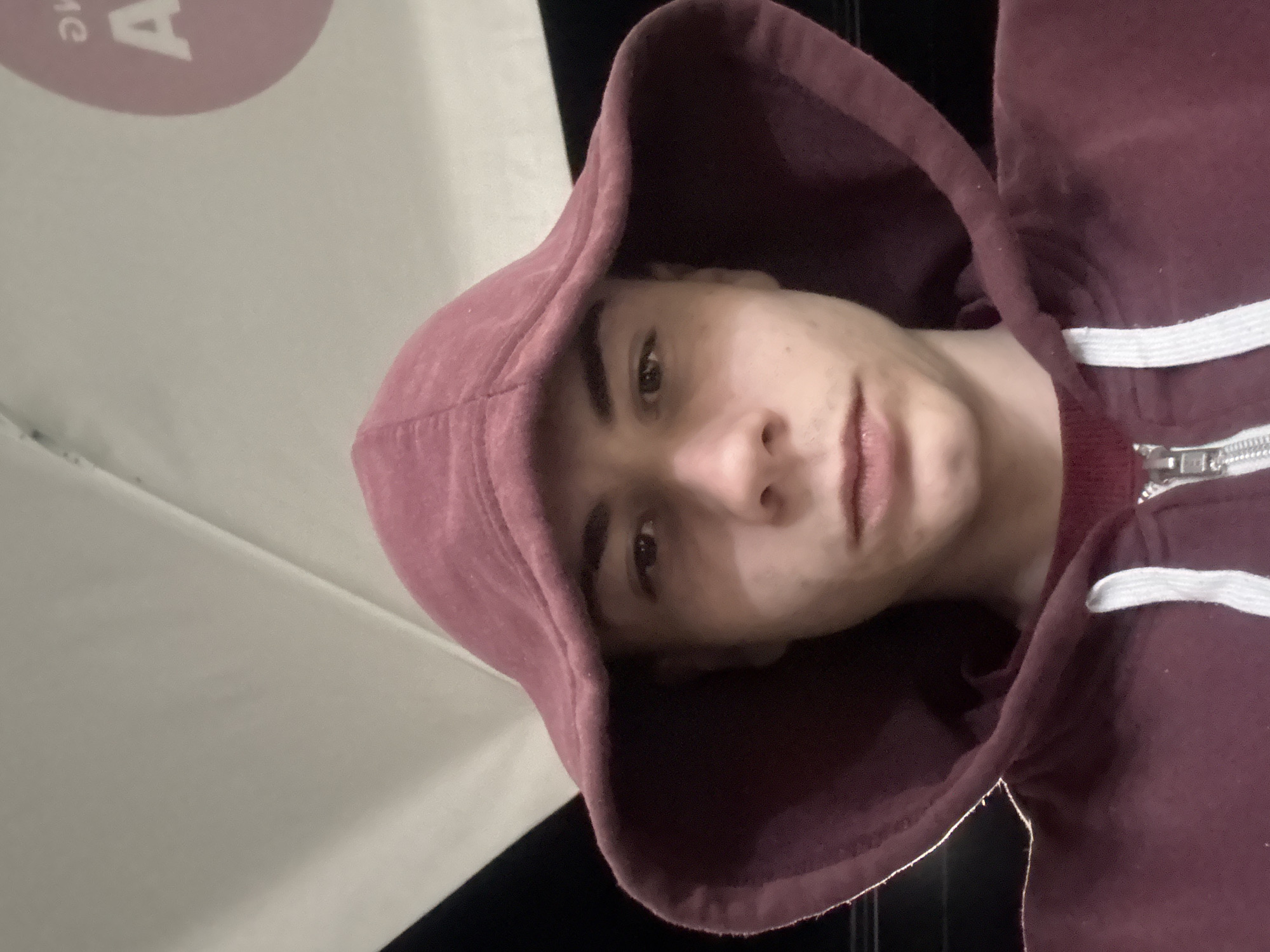A young person with light skin, wearing a maroon hoodie with white drawstrings and a zipper, faces the camera. They are under a partially visible white surface with a red logo on the left side. The background behind the white surface is black. The location cannot be determined from the image.