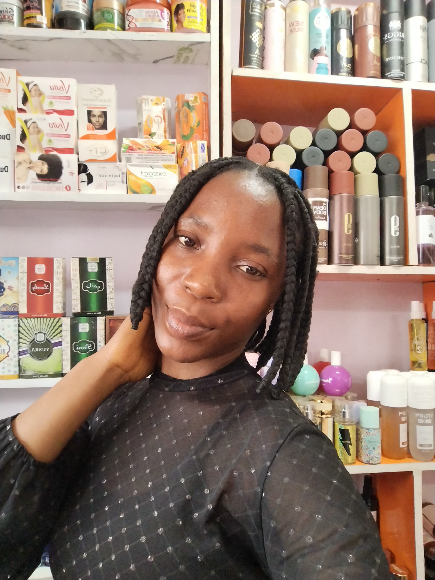 In Ugbowo, Nigeria, a young Black woman with braided hair is taking a selfie in what appears to be a small retail establishment selling beauty products. She is wearing a sheer black top with a diamond pattern. Behind her, shelves are filled with various cosmetic items, including soaps, lotions, perfumes, and body sprays. The products are arranged in an organized manner, with some boxes and bottles clearly visible. The lighting in the scene suggests it is daytime, and the environment seems to be an indoor space.
