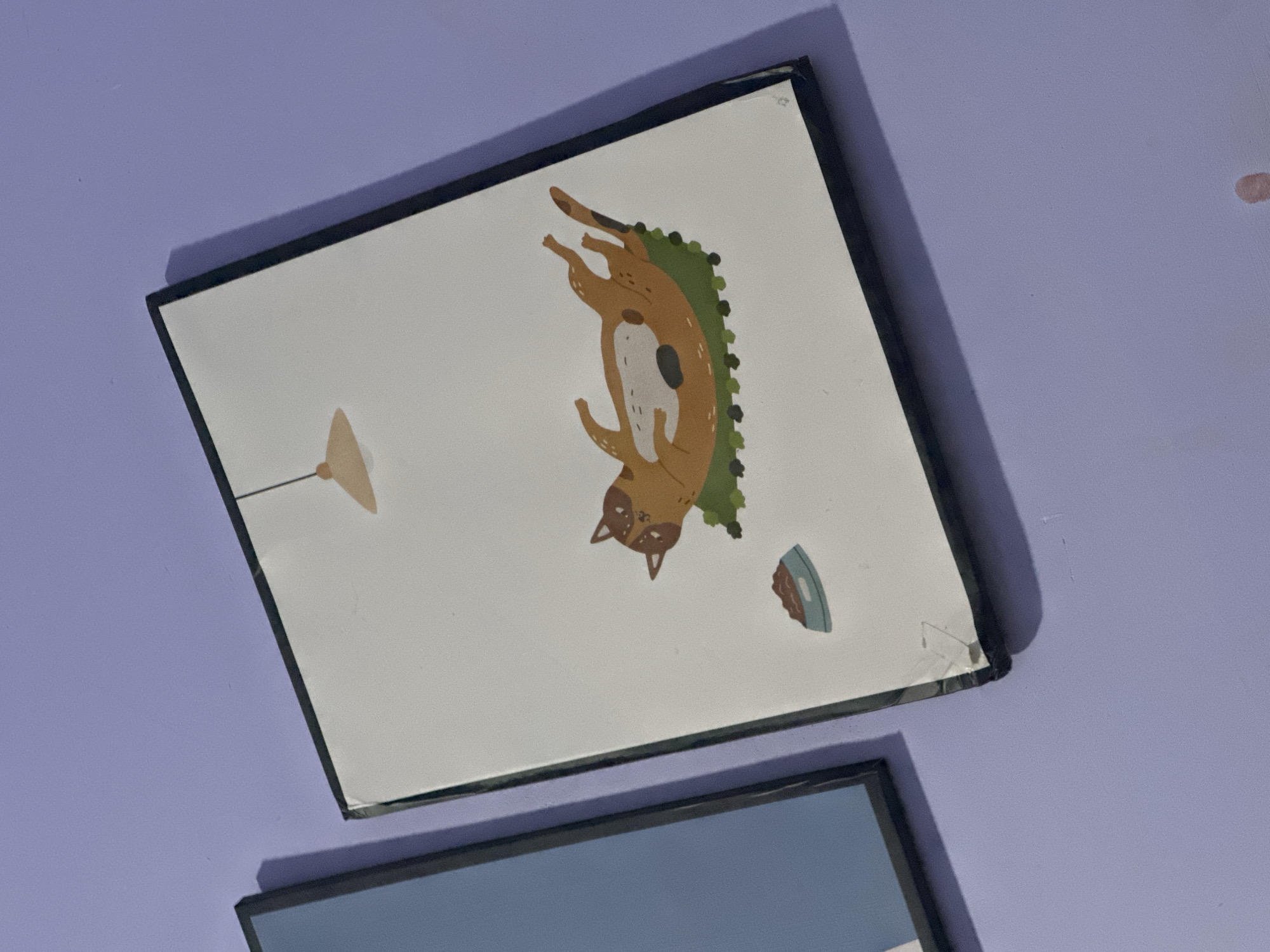 The image shows a close-up, eye-level view of two framed pictures hanging on a purple wall. The picture on the right is a white canvas with a cartoon illustration of a brown and white cat lying on its back on a green rug, with a bowl of food beside it. Above the cat, a simple beige lamp hangs from a thin black line. The frame of this picture is black and appears to be slightly damaged in the bottom right corner. The picture on the left is partially visible and has a blue and white design with a black frame. The wall is painted a light purple color and shows some subtle texture. Shadows are cast on the wall from the frames. The scene is indoors and appears to be a home or a small shop.