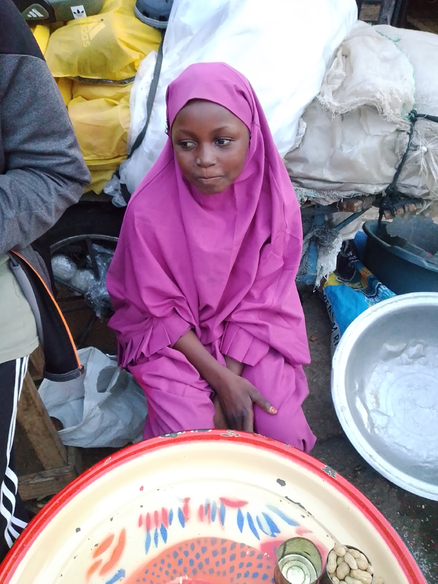 A young girl wearing a vibrant pink hijab and dress sits on the ground, looking to her left with a contemplative expression. She is positioned in what appears to be a marketplace or informal vendor setting. To her left, a person in a grey jacket and black pants with white stripes is partially visible, suggesting another individual present in the scene.
In the foreground, a large, colorful metal tray with a red rim is prominent. The tray is decorated with intricate patterns in red, blue, and orange. On the tray, there are two small metal cups; one appears empty, while the other is filled with peanuts.
Behind the girl, a collection of goods is piled up. These include yellow bags, some with visible "adidas" branding, and large white sacks, some of which are tied with rope. Other objects like a black bucket and a blue plastic bag are also visible amidst the merchandise. The ground is a mix of concrete and possibly dirt. The overall impression is one of a busy, everyday scene in a place where goods are being sold or distributed. The lighting suggests it could be daytime, though the specific time and weather conditions are not discernible from the image.