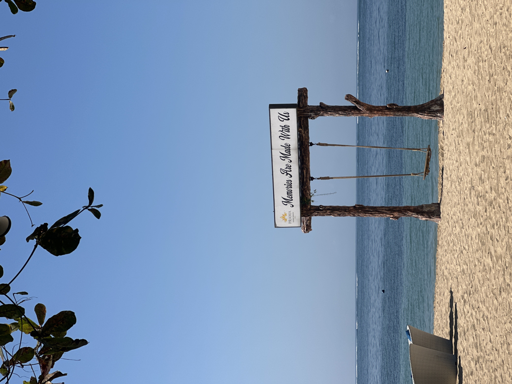 The image captures a serene beach scene in Denpasar, Indonesia, likely at a resort, as indicated by the "PRAMA SANUR BEACH HOTEL" logo on a sign. The foreground features a sandy beach with two white woven lounge chairs casting shadows. A rustic wooden swing set stands prominently, constructed from two thick, natural logs with a wooden plank swing suspended by ropes. Above the swing, a white sign reads "Memories Are Made With Us" in elegant script. The middle ground shows the calm, blue ocean stretching to the horizon, with subtle white wakes from distant boats. The sky is a clear, bright blue, and the top of the frame is partially obscured by the dark green leaves of a tree. The overall atmosphere is peaceful and inviting, perfect for relaxation and creating lasting memories.