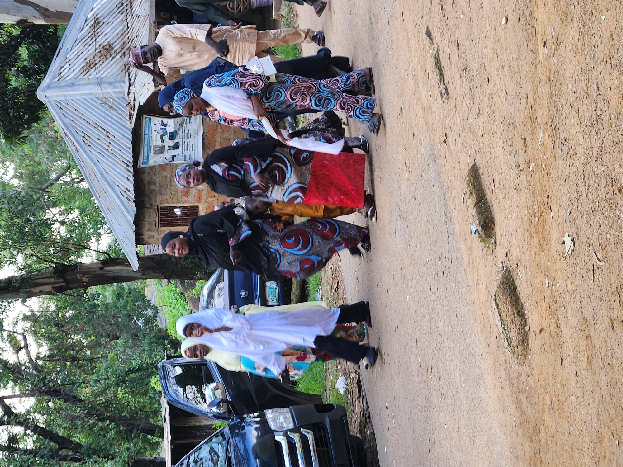 The image depicts a group of people, predominantly women, in what appears to be a rural or semi-urban setting in Jos, Nigeria. They are dressed in traditional African attire, with vibrant patterns and headscarves. Some individuals are wearing long, flowing dresses and trousers, while others are in more casual outfits. A few men are also present, one in a light-colored traditional garment and another in darker clothing.
The scene is set outdoors, with a dirt road in the foreground and a building with a corrugated metal roof visible in the background. Trees and lush green foliage suggest a tropical or sub-tropical environment. A dark-colored van, possibly a utility vehicle, is parked on the side of the road, with its doors open.
The people seem to be gathered for an event or a social occasion. Their expressions are neutral to slightly smiling, suggesting a calm or expectant atmosphere. The time of day is likely daytime, with soft, diffused light, hinting at an overcast or early morning/late afternoon period. The weather appears to be pleasant, with no signs of rain or extreme heat.
There is a poster or notice displayed on the wall of the building, featuring images and text, though the details are not legible. One of the women is holding a white cloth, and another is holding a red object, possibly a gift or religious item. The overall impression is one of community gathering and cultural tradition.
