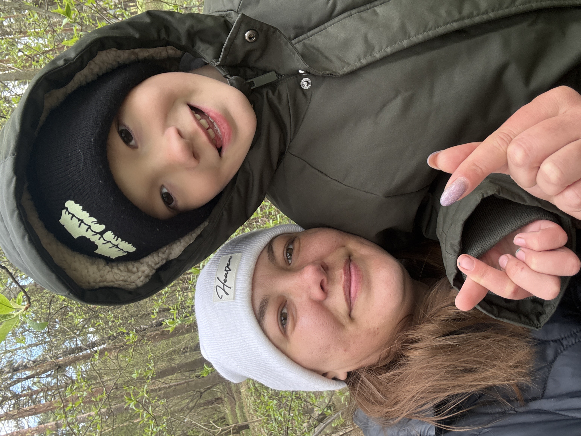A rotated medium shot depicts a woman and child outdoors, potentially in a park or wooded area. The woman, wearing a light-colored beanie with a visible "Heaven" tag, is positioned at the bottom of the frame. She is looking at the camera and smiling. A child, possibly her son, is above her, wearing a black beanie and a green hooded jacket with a light-colored fur lining. The child is also smiling and looking at the camera. A hand is visible, making a mini-heart gesture with the fingers. Trees and greenery are visible in the background. While the city is not recognizable, the general atmosphere is consistent with an outdoor scene in Dmitrov, Russia.