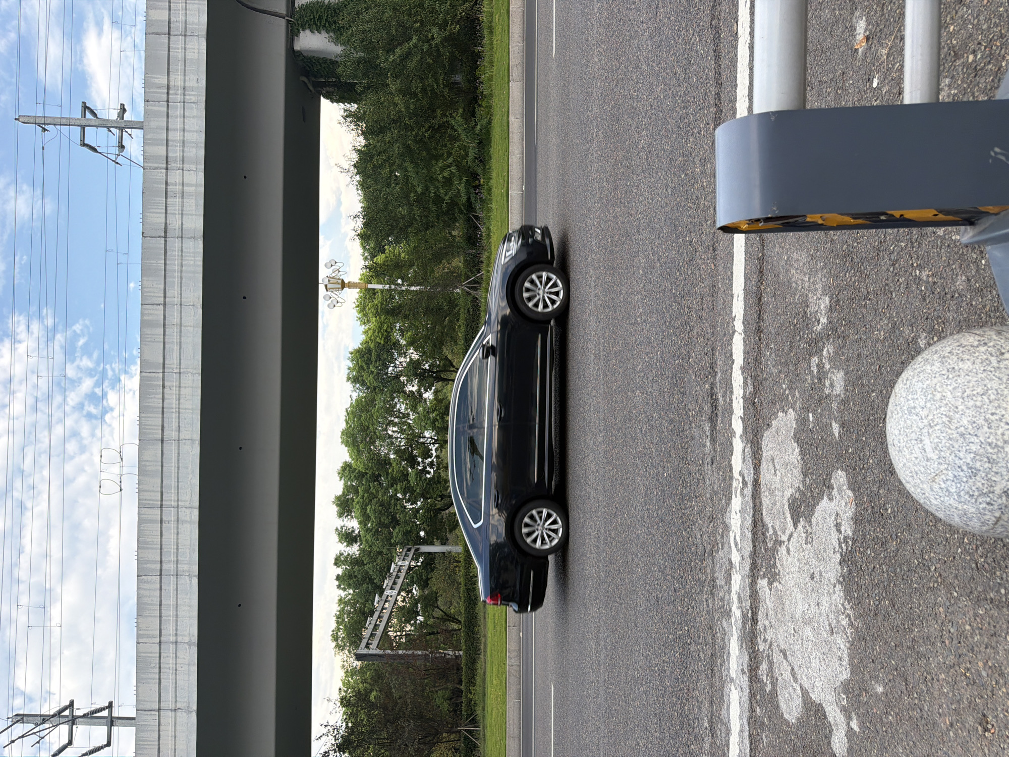 In Jiangjin, China, a black sedan is captured in motion on a paved road, driving beneath a large concrete overpass. The overpass is supported by pillars and has overhead power lines. Trees and greenery line the side of the road, with a lamppost visible in the background. The sky above is partially cloudy. In the foreground, a white line is painted on the asphalt, and a segment of a grey metal barrier with yellow detailing is visible.