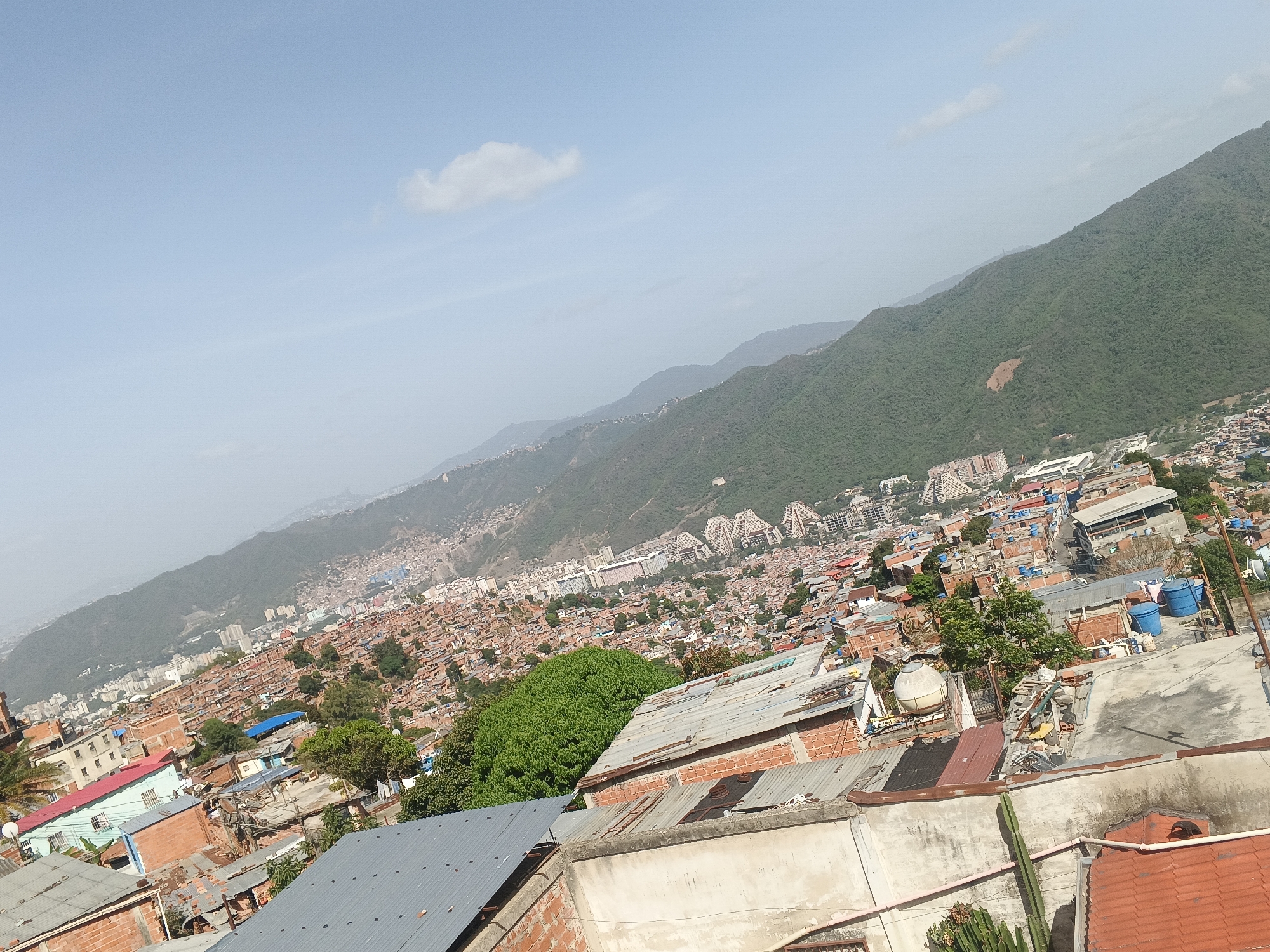 The image depicts a densely populated hillside neighborhood in Caracas, Venezuela, viewed from an elevated perspective. The foreground shows rooftops constructed from corrugated metal and brick. The midground reveals a sprawling urban landscape of closely packed dwellings, punctuated by patches of trees. In the distance, a large mountain range dominates the skyline. The sky is pale blue with a single cloud. The photograph conveys a sense of dense urban development juxtaposed against a natural landscape.