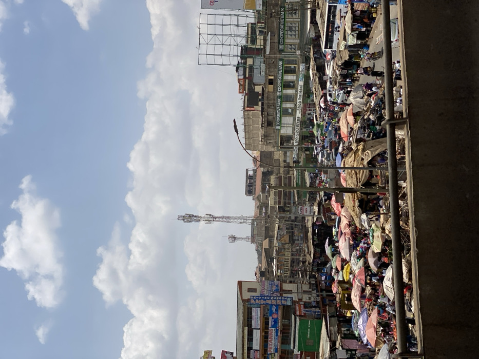 The image shows a busy street market in Githurai, Kenya, taken from a high vantage point. The view looks down upon a bustling market scene, with numerous vendors selling goods under brightly colored tarpaulins and umbrellas. The image is taken from an elevated location, likely a bridge or walkway, providing a bird's-eye view of the market and surrounding buildings. The foreground is dominated by a cement wall, which partially obscures the market activity. The market extends back to the left, where it meets a series of multi-story buildings. In the background, a cell phone tower is visible against a partly cloudy sky. The buildings on either side of the market display various signs, including those for Cooperative Bank and a shop for sale. There are many people in the market, walking between stalls, haggling with vendors, or carrying goods. The scene is characterized by a sense of organized chaos, with the colors of the umbrellas and merchandise contrasting with the gray concrete of the buildings and the bright blue sky. The overall impression is one of a vibrant and energetic commercial hub.