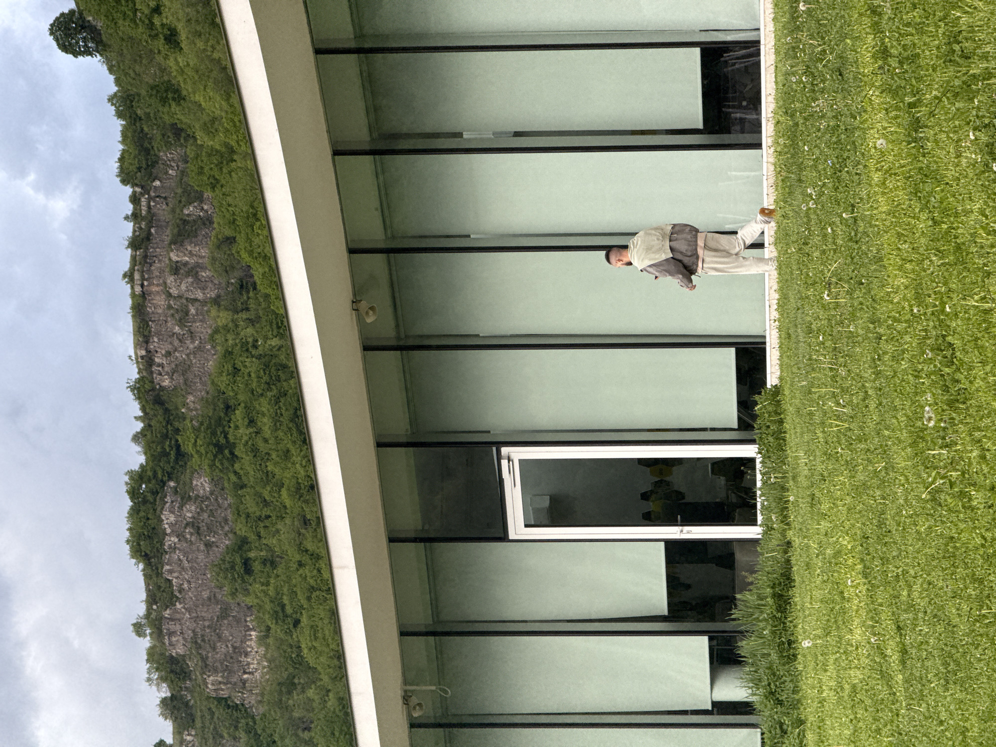 The image shows a man walking on grass in front of a modern building with large windows, located in Chkalov, Armenia. A rocky, green cliff is visible to the left of the building. The sky is overcast. The man is wearing light-colored pants, a light shirt, and a darker jacket. There is a security camera mounted on the building's exterior.