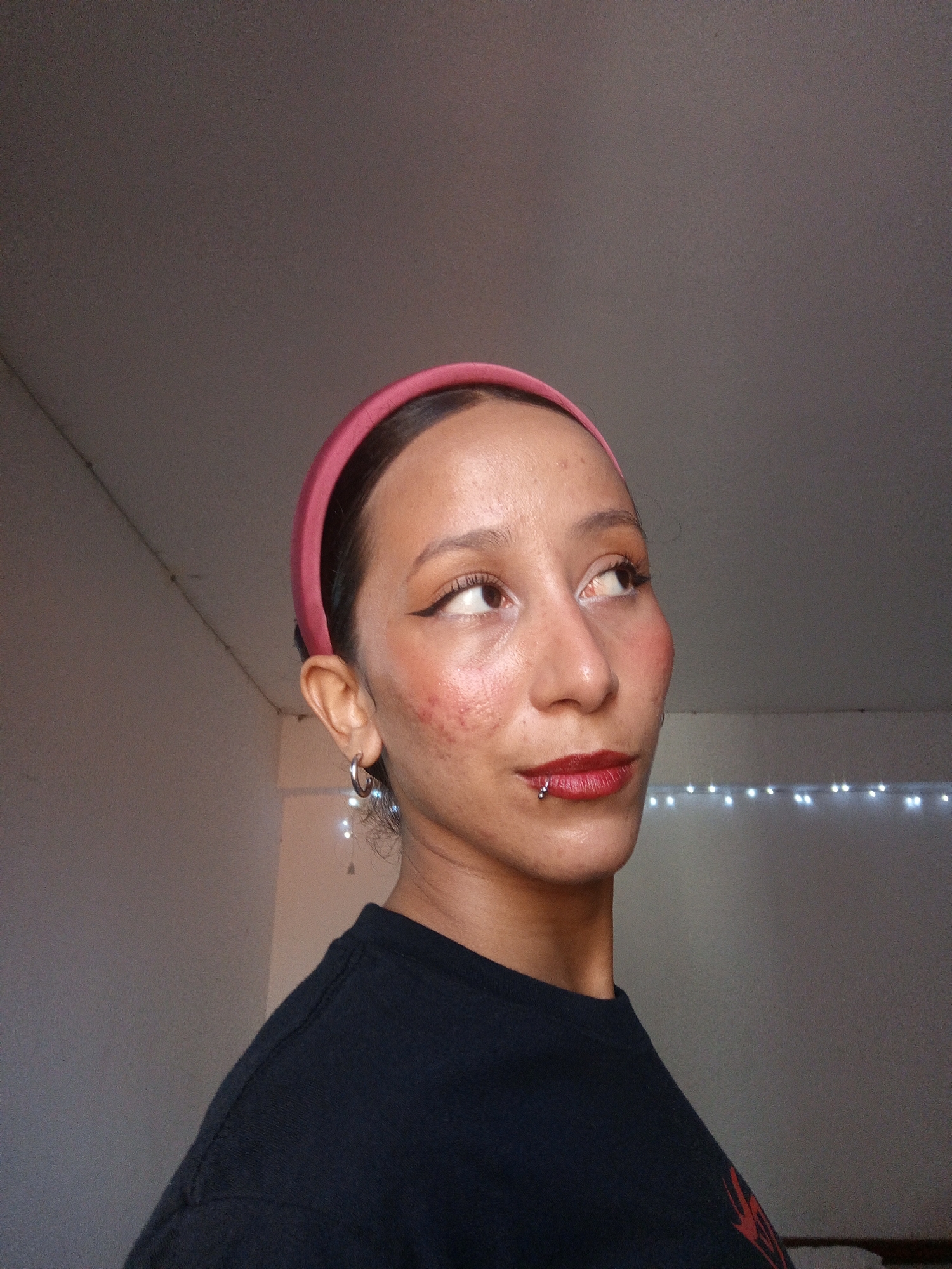 The image shows a woman with light brown skin wearing a pink headband and a black t-shirt. She has dark eyeliner and red lipstick, with a lip piercing on the lower lip. She is looking upwards and to the right. She has hoop earrings, and what appears to be a string of lights can be seen in the background on a wall. The location in the city of Porlamar, Venezuela, cannot be determined from the provided image.