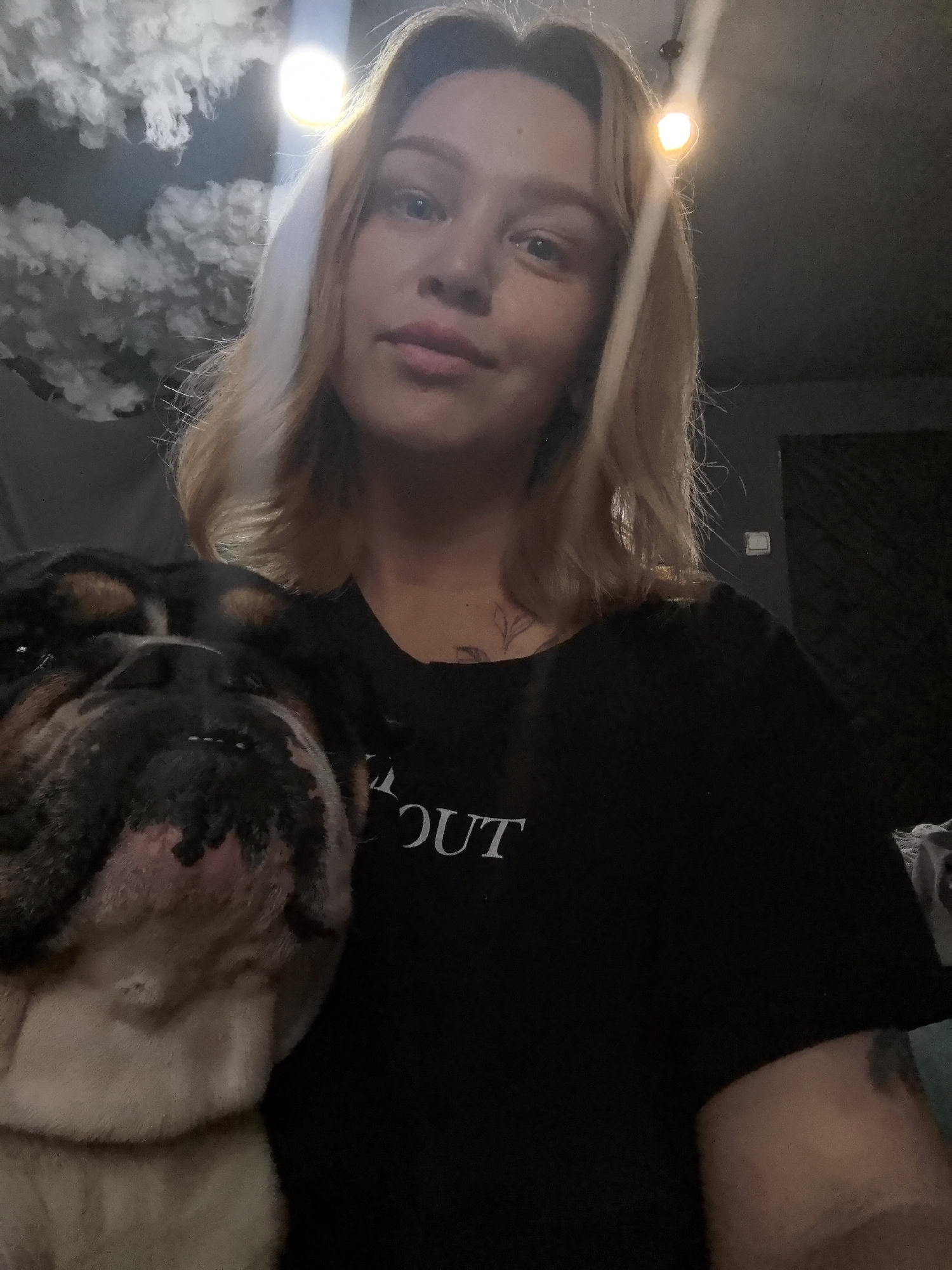 A person with blonde hair and light skin is shown from the chest up, facing the camera. They are wearing a black t-shirt with white lettering, partially obscured by the presence of a bulldog's head in the foreground. The dog is mostly white and black, with a wrinkled face. The background is dark and includes a lighting fixture, potentially indicating an indoor setting. There are fluffy white shapes above the person that look like clouds. A light switch is visible in the background. There is no information to confirm the location to be Noviy, Russia.