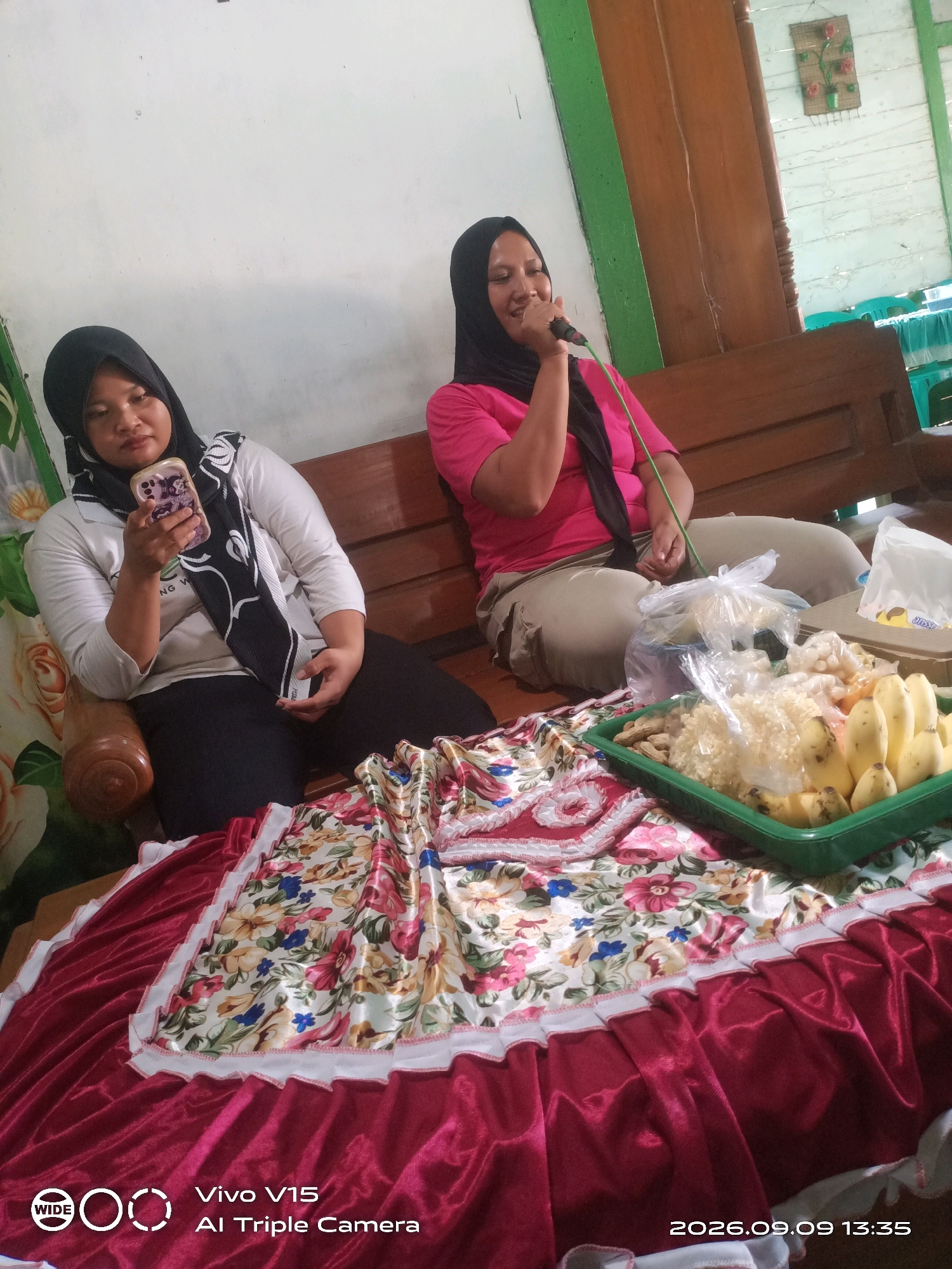 In a room with white walls, two women are seated on a wooden bench. The woman on the left, wearing a black hijab and white shirt, holds a mobile phone. The woman on the right, also wearing a black hijab, is dressed in a pink t-shirt and beige pants, and is speaking into a microphone. In front of them, on a table covered with a floral and red fabric, is a green tray filled with bananas and other food items in plastic bags. The overall setting appears to be a domestic or community gathering space in Plosorejo, Indonesia, indicated by the text watermark "Vivo V15 Al Triple Camera" and the date "2026.09.09 13:35".