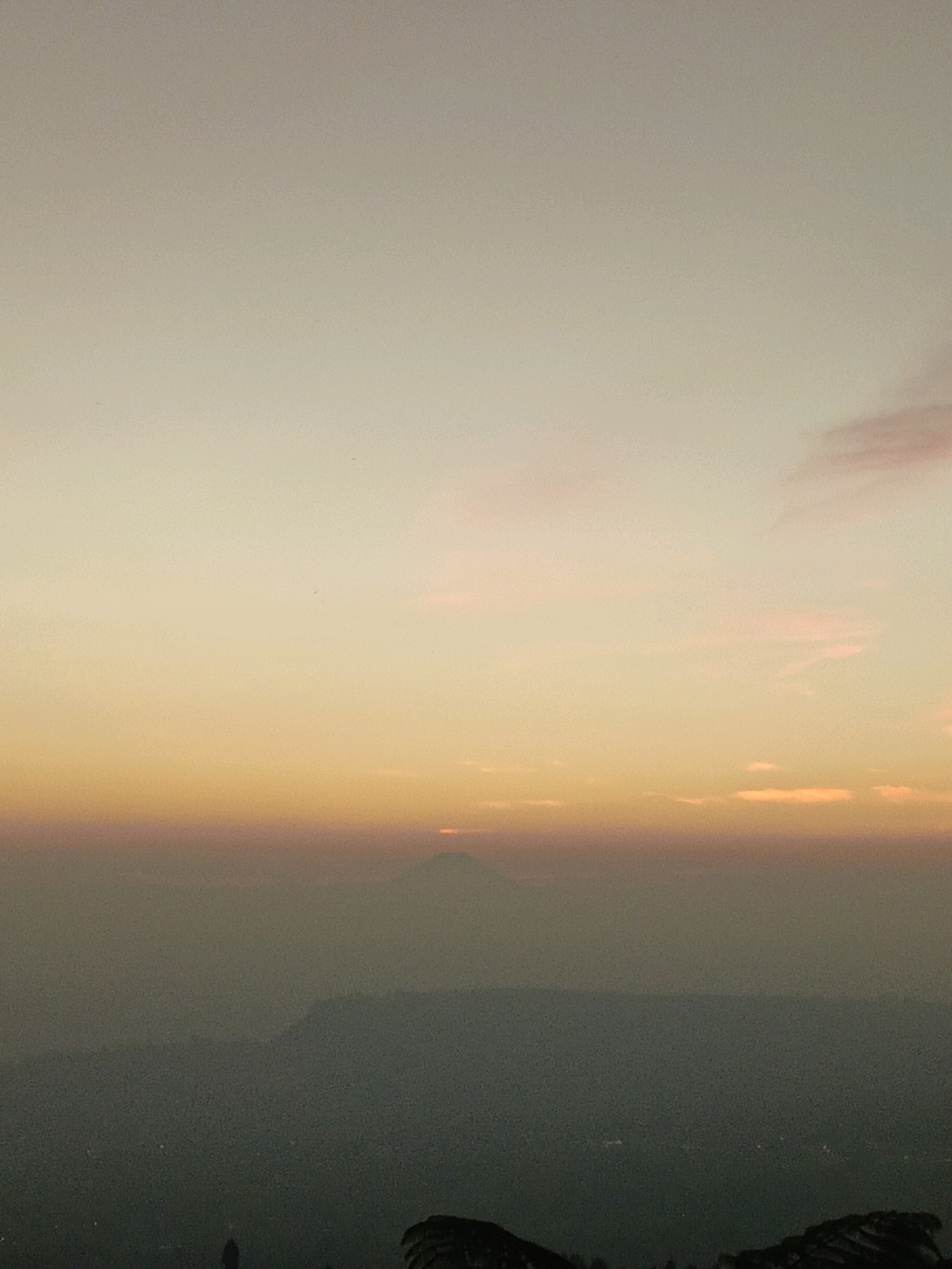 An atmospheric landscape depicts a hazy sunset over a mountainous region, likely in Butuh Kidul, Indonesia. The sky transitions from a pale yellow at the top to a soft orange and pink gradient closer to the horizon. In the distance, a distinct mountain peak, possibly Mount Merapi, is visible through the haze, with a small, bright light at its summit suggesting the setting sun. Below the horizon line, layers of subdued, dark green hills fade into the foreground, with faint outlines of what might be a village or scattered lights visible in the valley. The foreground is dominated by silhouetted, dark, rocky formations, with a lone, dark human figure standing in the lower left portion of the frame. The overall impression is one of serene, natural beauty, enhanced by the muted colors and diffused light.