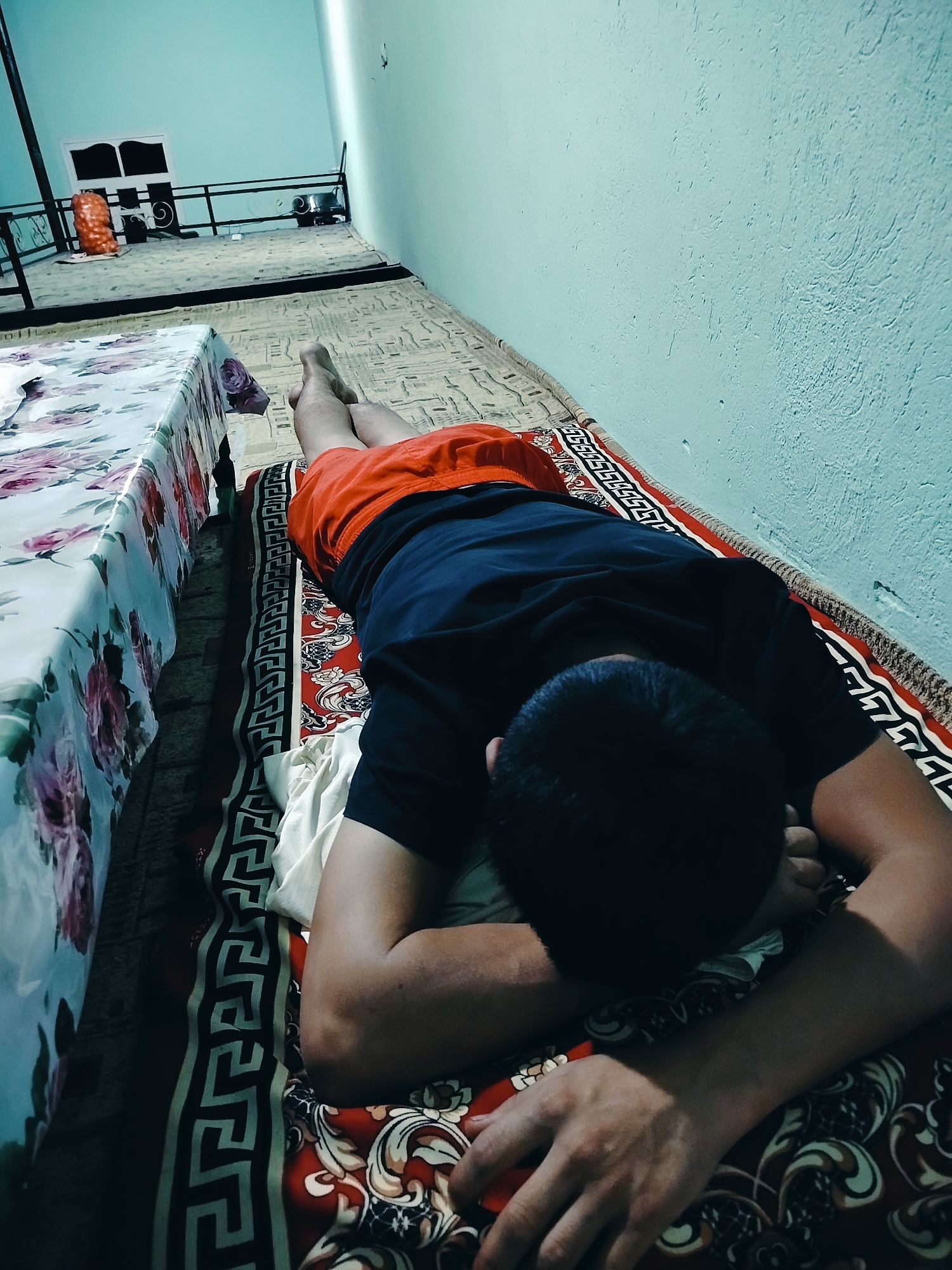 A young person lies on their side on a patterned rug on the floor. They are wearing a dark blue t-shirt and bright orange shorts. Their head is resting on their arms, which are bent at the elbows. One leg is extended, while the other is bent at the knee. To the left of the person, a table covered with a floral cloth is visible. The room has light blue walls. In the background, a window or opening looks out onto what appears to be an outdoor balcony or rooftop area with railings, a bag of what looks like onions, and some electronic equipment. The overall scene suggests a casual, indoor setting, possibly a bedroom or living area. The image was taken in the city of Kölkent, Kazakhstan.