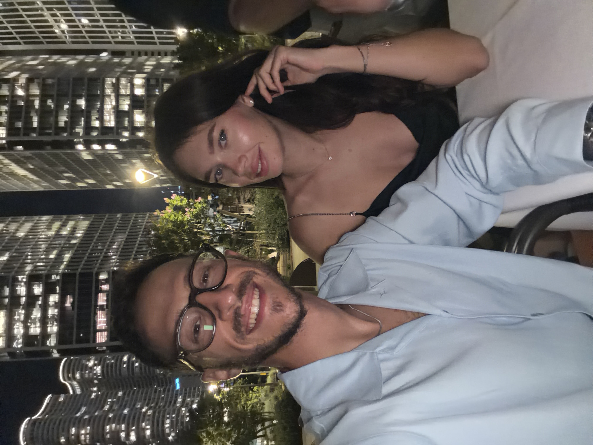 A selfie taken at night in Tel Aviv, Israel, captures a smiling man in the foreground wearing glasses and a light blue button-down shirt, with a woman in a dark dress to his right. Behind them, illuminated skyscrapers and trees form the urban backdrop. The man's left arm is on a table, while the woman rests her head on her left hand, touching her right ear.