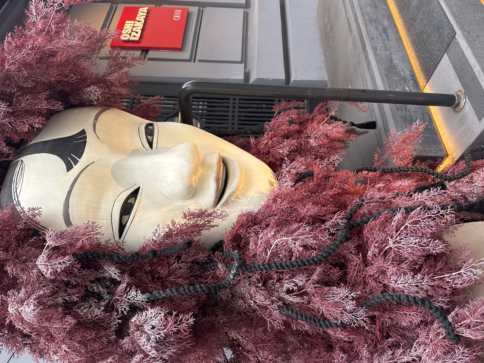 A large, light-colored mask or sculpture of a human face dominates the foreground, lying horizontally amidst a dense bed of dark pink and reddish artificial foliage. The mask features elongated, serene eyes and distinctive black painted eyebrows and eye details, evoking traditional Asian mask aesthetics. Black braided cords are artfully intertwined within the intricate, coral-like foliage.
The display is part of an outdoor or semi-outdoor installation, likely decorating the facade of a building in Moscow, Russia. In the background, a bright red sign with white lettering prominently displays "OSHI IZAKAYA" and a smaller logo featuring a character, possibly "B," within a square.
Surrounding the central display are grey architectural elements, including textured walls and what appears to be a grated vent. A black metal bar extends horizontally, possibly acting as a railing or structural support. To the right, a section of the wall features a warm yellow light strip, hinting at a decorative lighting scheme. The scene is brightly illuminated, indicating daytime. The photograph is captured at an oblique angle, presenting a dramatic and decorative art piece.
