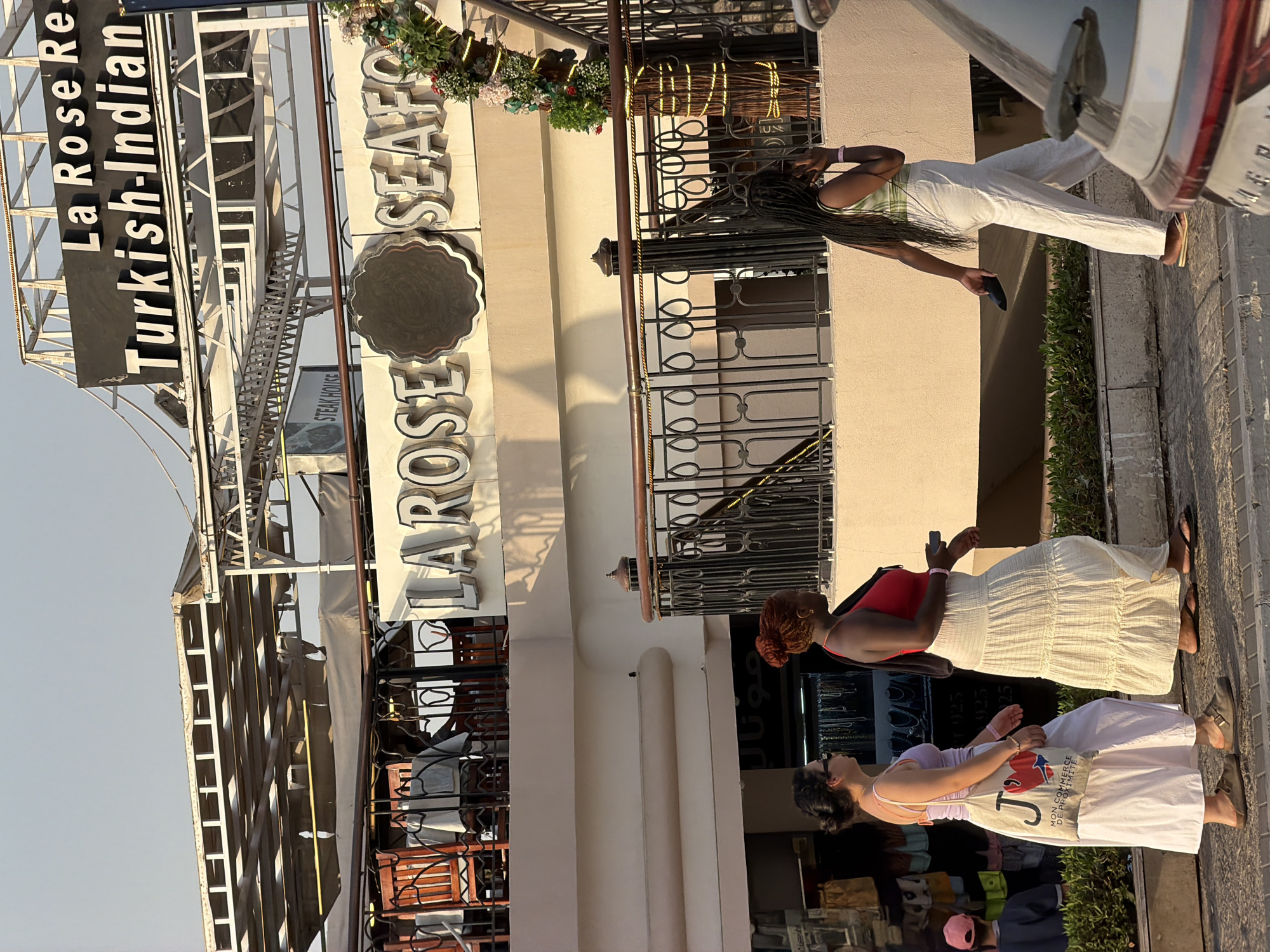 In Sharm El Sheikh, Egypt, three women are walking along a paved walkway in front of a multi-story building. The building features signage for "La Rose Rest. Turkish-Indian" and "LAROSE SEAFLOOR" and other businesses on upper levels. The woman on the left wears a light purple tank top, white pants, and carries a tote bag. The woman in the middle is in a red tank top and a long, cream-colored skirt. The woman on the right has braided hair and is wearing a green and white striped top and white pants, holding a dark object and a mobile phone. The scene is illuminated by natural sunlight.