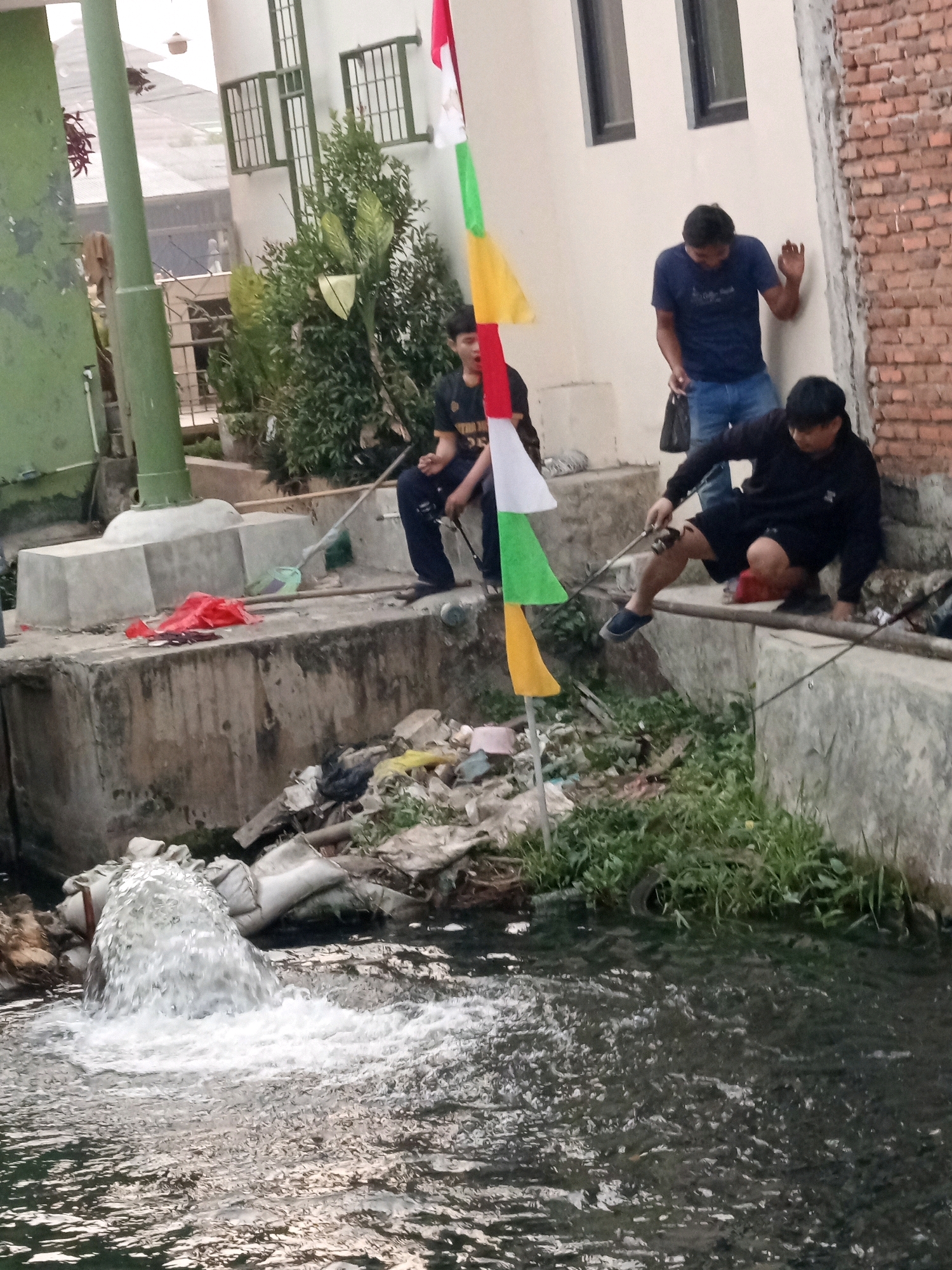 The image depicts an outdoor scene in Karawang, Indonesia, where three individuals are engaged in fishing activity by a canal. The canal's water is visibly polluted and overflowing from a pipe, suggesting a drainage or waste discharge. The environment is characterized by urban elements such as brick buildings and concrete structures, juxtaposed with overgrown vegetation and scattered debris along the canal banks.
In the foreground, a young person in dark clothing is seated on the edge of the canal, holding a fishing rod. To their left, another young person is also seated and appears to be fishing, with a fishing rod in hand. Further to the right, an adult male in a dark blue t-shirt and jeans stands near a brick wall, holding a dark bag and seemingly observing the others. A colorful flag, composed of red, white, green, and yellow sections, stands planted in the foreground, adding a splash of color to the otherwise utilitarian setting. The overall impression is one of a community engaged in a recreational activity amidst a less-than-ideal urban environment, highlighting the presence of both human activity and environmental challenges.
