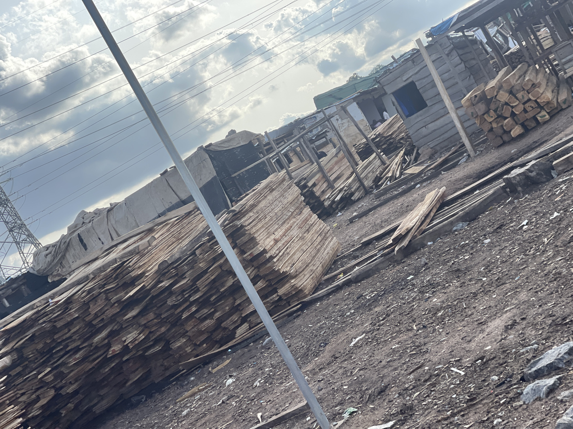 The image depicts a lumberyard or wood storage area located in what appears to be a densely populated or informal settlement in Abuja, Nigeria. Large stacks of wooden planks dominate the foreground, with various lengths and thicknesses visible. The planks are stacked irregularly.
In the background, simple wooden structures or shelters are visible, likely serving as workshops or storage for the lumberyard. Some people can be seen in the background, near the structures.
The ground is dirt and gravel. An electrical transmission tower is on the left. The sky is overcast.
