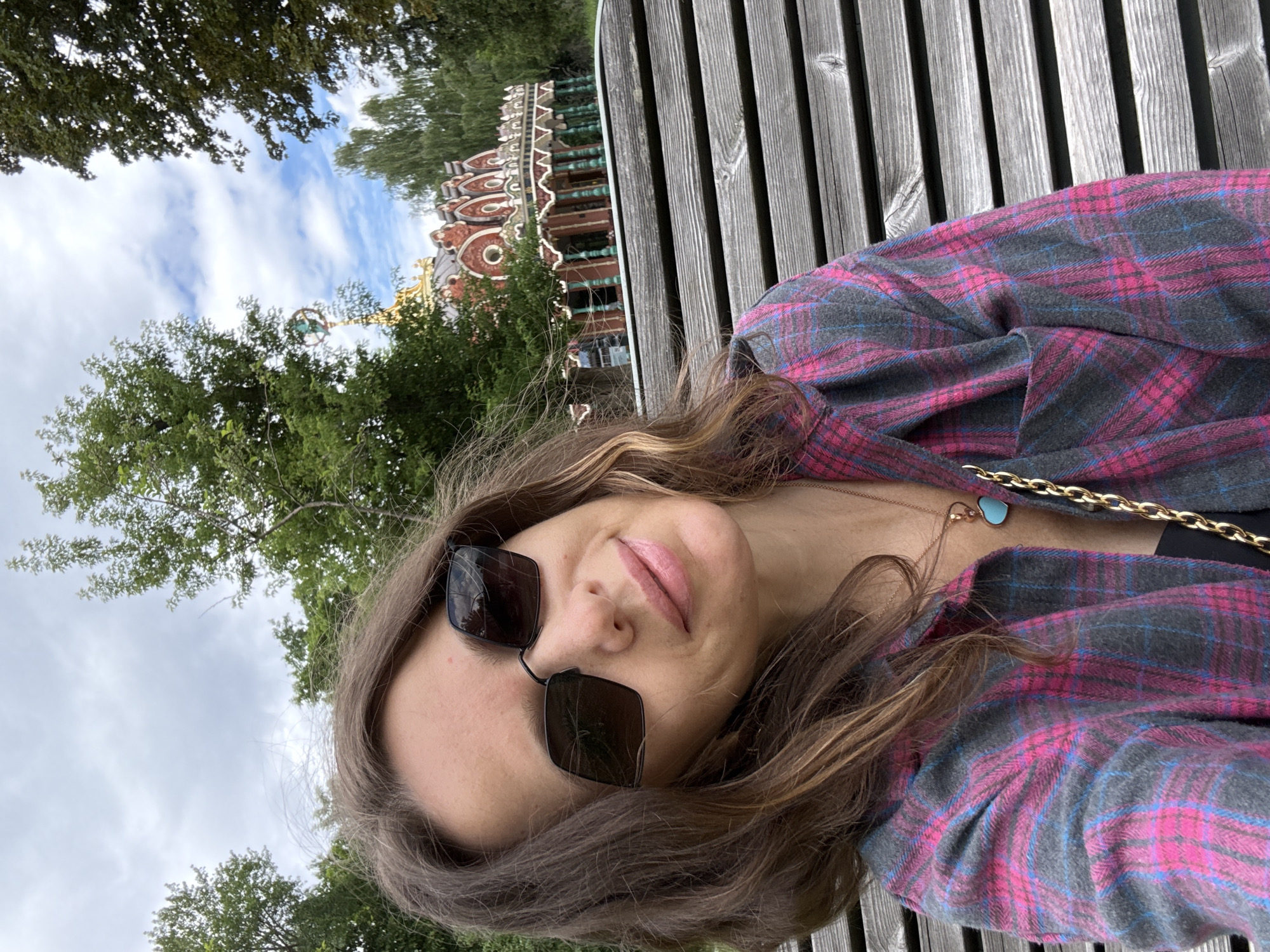 A medium shot captures a woman from the chest up, seated on a wooden slatted bench. She is wearing a pink and grey plaid shirt and sunglasses, with a gold chain necklace and a blue heart-shaped pendant. Her hair is brown and falls over her shoulders. In the background, a red and gold ornate building with arched windows and pillars is visible amidst lush green trees. The sky is partly cloudy. This appears to be the Izmaylovo Kremlin, a Russian architectural complex in Moscow.