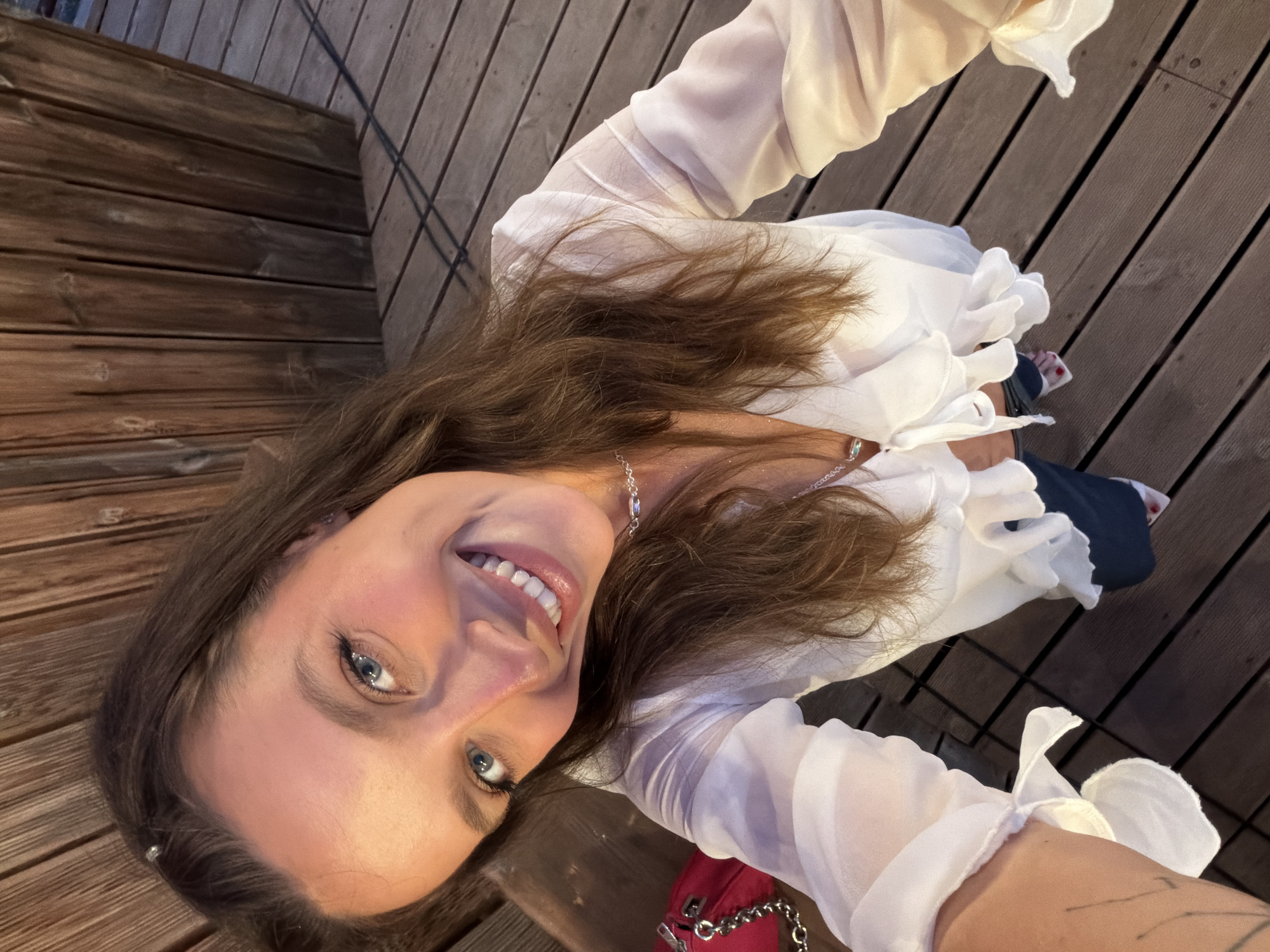 A high-angle selfie captures a woman with shoulder-length brown hair, fair skin, and blue eyes. She is smiling broadly. She wears a sheer, white blouse with ruffled detailing. A silver necklace adorns her neck. Part of a red handbag with a silver chain strap is visible at the bottom of the frame. She appears to be seated on a wooden structure, possibly a bench or deck, composed of evenly spaced planks. The location may be Bigova, Montenegro.