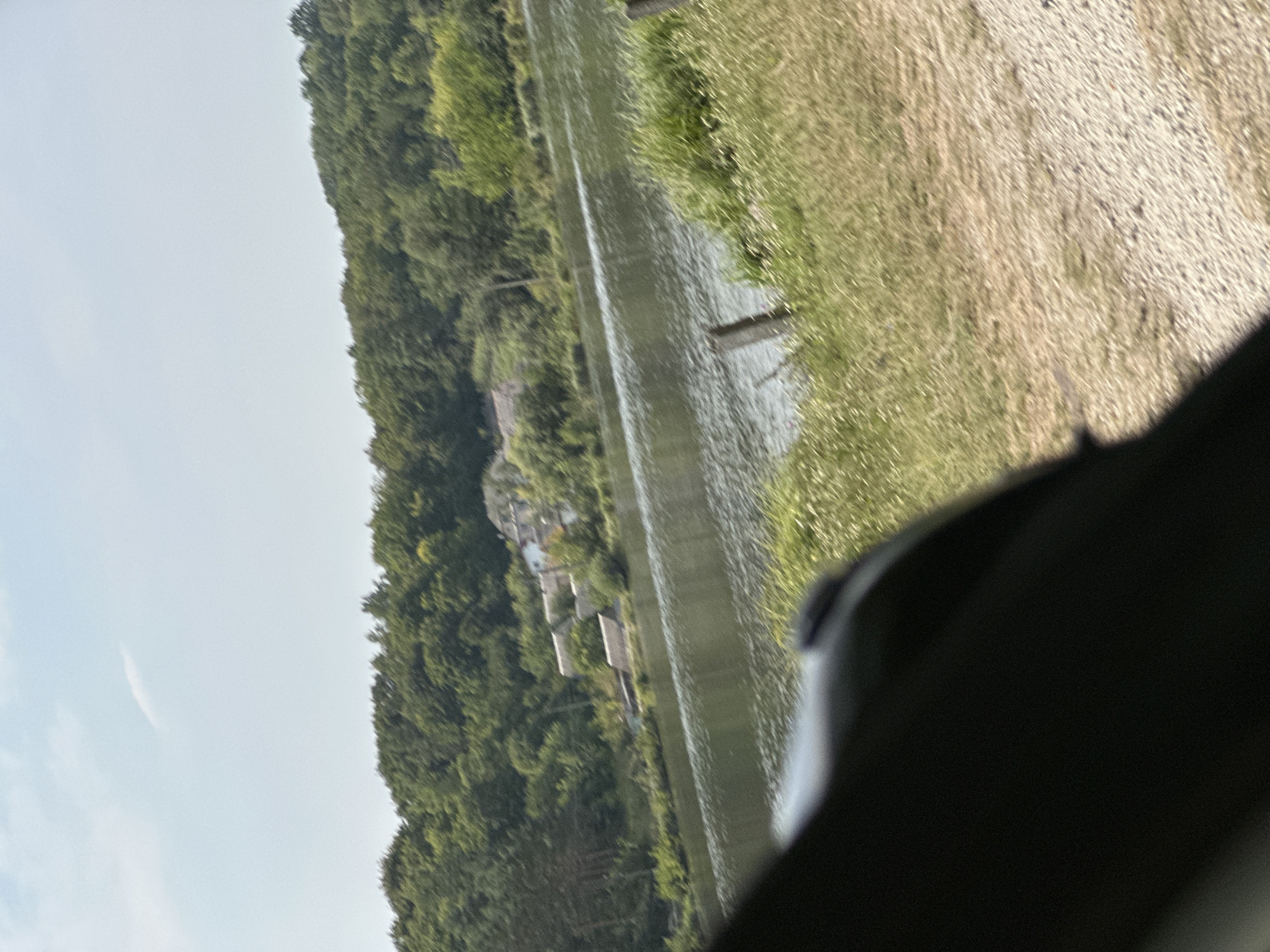 This is a slightly blurry, wide-angle photograph taken from a low vantage point, possibly from within a vehicle, looking out at a natural landscape. The foreground is dominated by a dark, indistinct object which obscures the lower left portion of the frame. To the right of this object, the ground appears to be a mixture of gravel and dry grass, with some taller, green grass and thin, wooden posts.
Beyond this foreground, a body of water, likely a lake or river, stretches across the middle ground of the image. The water's surface is rippled and reflects the light, creating a shimmering effect. On the far side of the water, a dense, green forest covers a hillside. Within the trees, a cluster of buildings, possibly houses or cottages, are visible, some with visible roofs. The sky above is a pale blue with a few wispy white clouds.
The overall impression is a serene, rural scene on a sunny day. The image has been identified as being located in or near the village of Makiv, Ukraine.