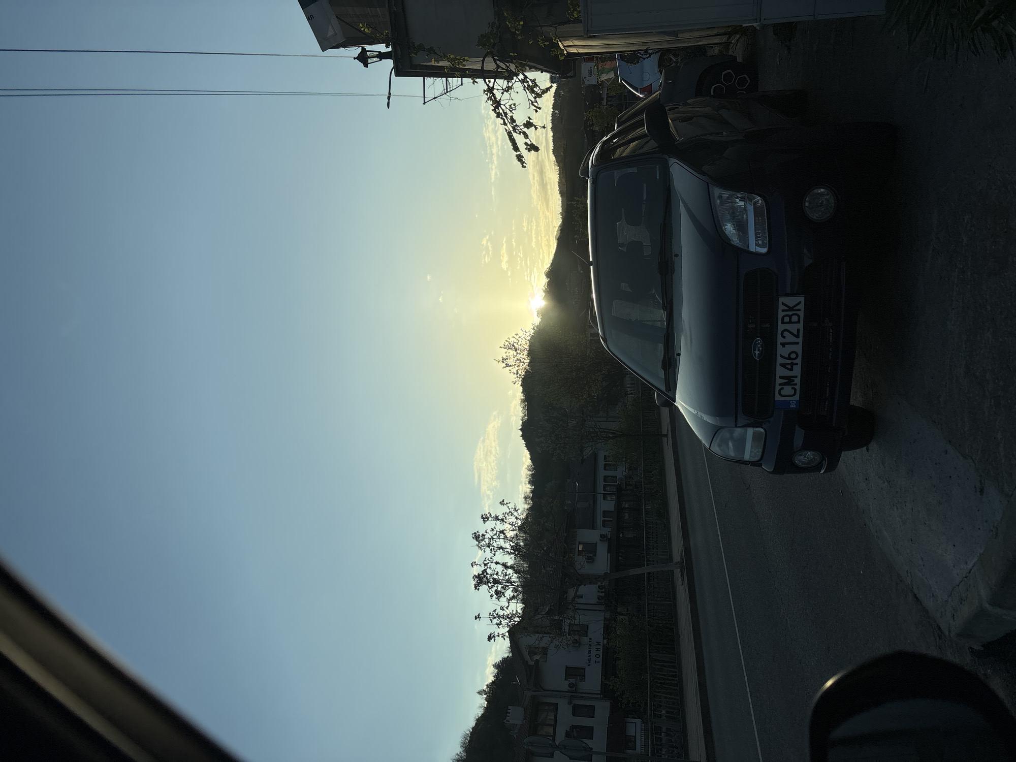 The image, originally rotated, captures a serene outdoor scene at either sunrise or sunset, viewed from inside a vehicle. The lighting suggests late afternoon or early morning, with the sun low on the horizon, casting a bright glow over a distant mountain range and parts of the sky.
In the foreground, the lower left corner shows dark, blurred elements, likely the interior frame or dashboard of the car from which the photo was taken.
To the right, a dark blue or black Subaru SUV or crossover is prominently parked on the side of a paved road. Its front fascia, headlights, and grille are visible, along with a Bulgarian license plate displaying "CM 4612 BK". The "CM" prefix indicates the Smolyan region of Bulgaria, consistent with the specified location context of Zlatograd.
The road runs towards the background, flanked by buildings and trees. On the left side of the road, several white buildings, resembling houses or small hotels, are visible among trees with sparse foliage. One of these buildings clearly displays Cyrillic text, which, when rotated and mirrored, reads "ХОТЕЛ-РЕСТОРАНТ ТОНИ" (HOTEL-RESTAURANT TONI).
In the background, a large hill or mountain range is silhouetted against the bright sky, with the sun appearing directly behind it, creating a starburst effect and warm illumination across the horizon. The sky is predominantly clear blue, with a few soft, wispy clouds reflecting the golden light. Utility lines are visible in the upper left portion of the image. The overall impression is one of tranquility in a small town or rural setting.