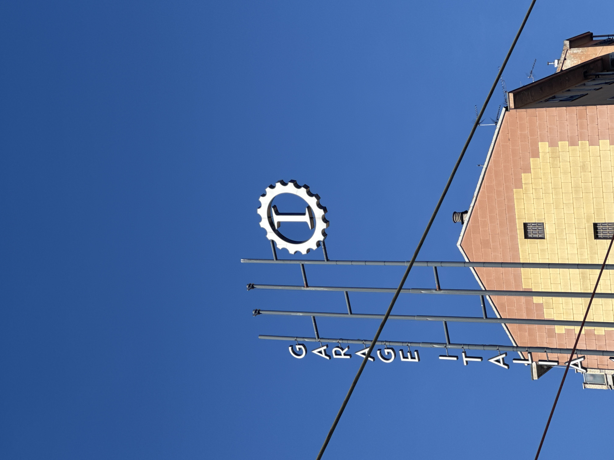 A low-angle shot reveals a sign reading "GARAGE ITALIA," topped with a gear-shaped logo containing a prominent "I." The sign is suspended in front of a building with a patterned facade of alternating peach and yellow tiles. Electrical wires cut across the sky, which is a clear, deep blue. The location is Milan, Italy, as indicated by the sign's name, "Garage Italia," which is a notable establishment in the city.