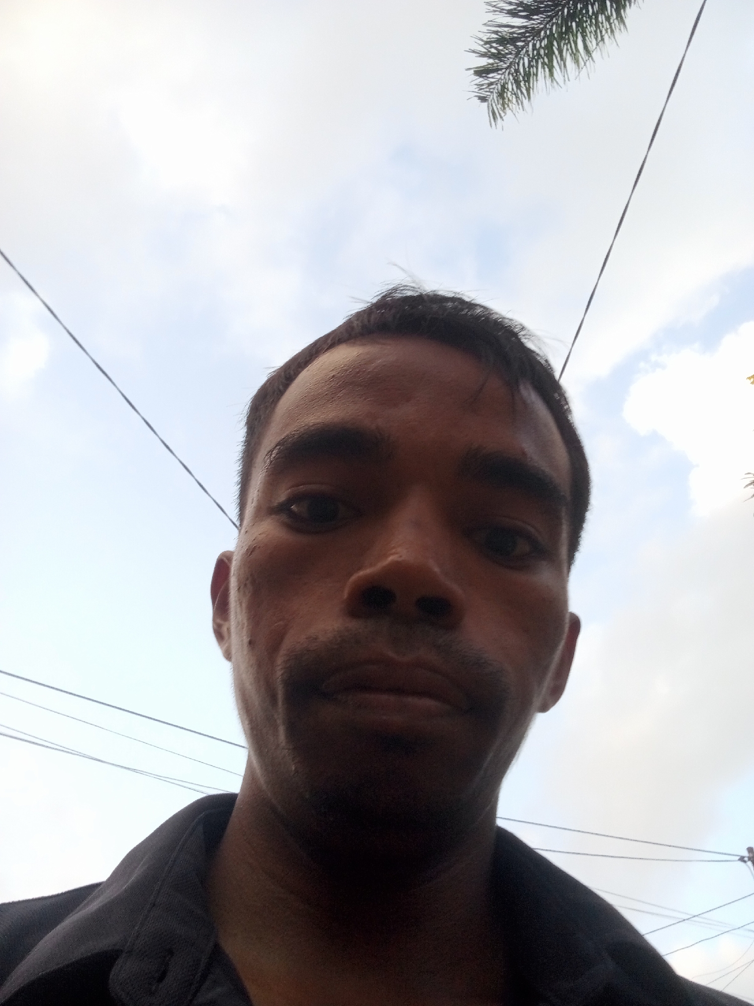 A low-angle, close-up selfie of a man facing the camera, taken outdoors in Surabaya, Indonesia. The man has dark skin, short dark hair, and is wearing a dark collared shirt. The background consists of a cloudy sky with power lines and some foliage visible at the top. The overall lighting suggests it is daytime.