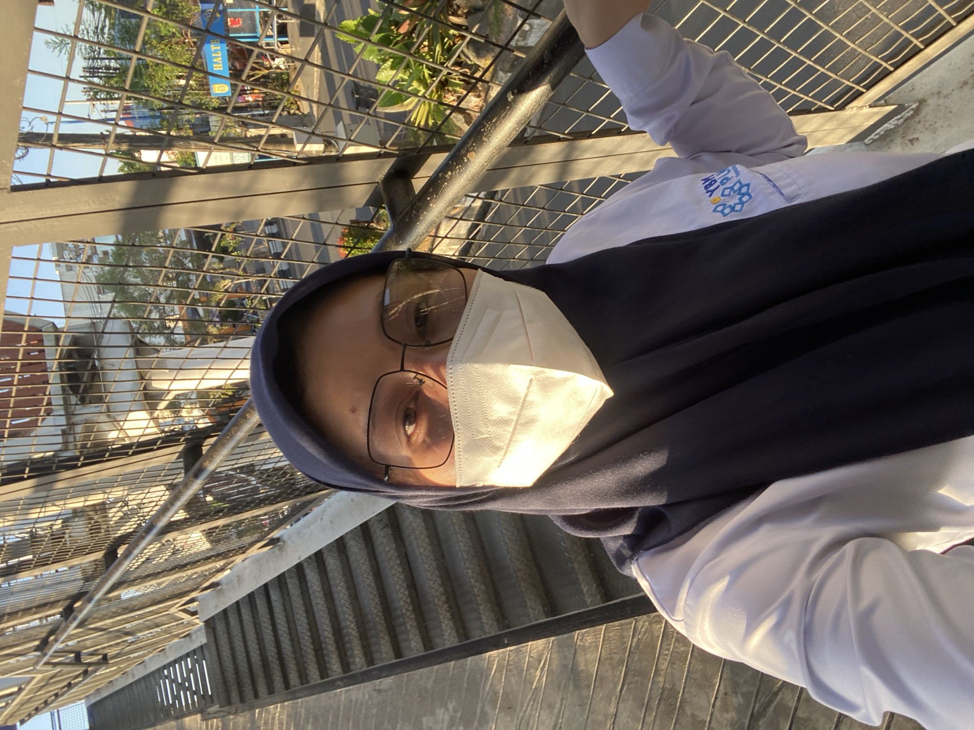 A woman wearing a dark blue hijab, glasses, and a white face mask takes a selfie on a pedestrian overpass in Banjarbaru, Indonesia. She is wearing a white long-sleeved shirt with a blue and white logo on the left breast. The overpass has metal stairs and railings. In the background, there is a street with traffic and buildings, including a blue sign that reads "HALTE." The sun is shining, casting long shadows.