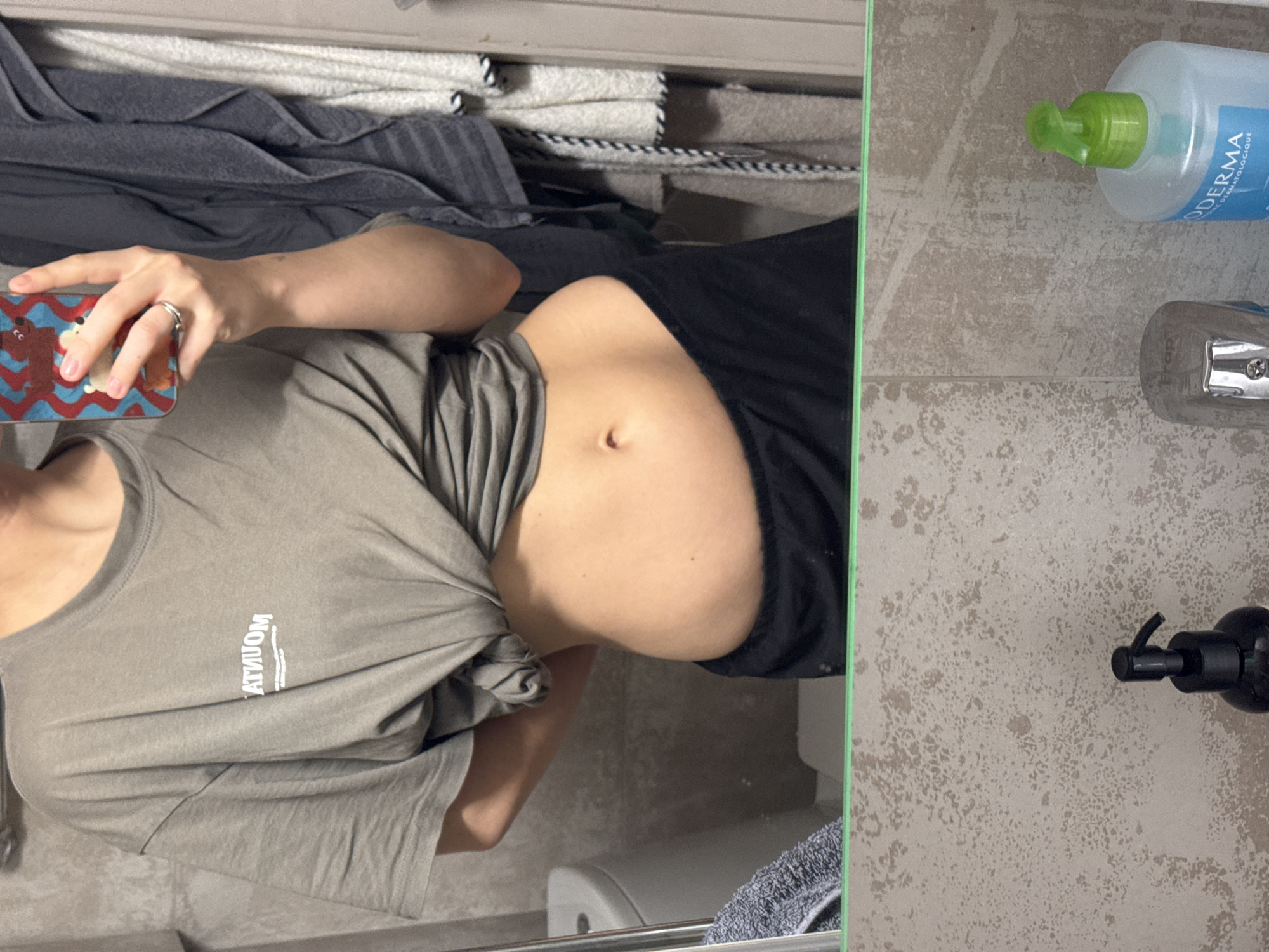 This indoor mirror selfie captures a person's torso, left arm, and hand, taken in what appears to be a bathroom or dressing area. The individual, seen from the mid-chest down, wears a loose, light brown or grey t-shirt pulled up to reveal a slightly rounded belly. Dark shorts or underwear are partially visible on the right side of the midsection. A silver-colored ring adorns the ring finger of the left hand, which holds a smartphone encased in a distinctive blue and red patterned cover featuring cartoon-like animal figures.
In the reflected background, neatly folded grey and white striped towels are stacked on a shelf. A white porcelain object, possibly a toilet or bidet, is partly visible in the lower left of the reflection. On the right side of the image, outside the mirror's frame, two pump dispenser bottles are placed on a textured, light-colored surface resembling bathroom tiles. One is a light blue bottle with a green pump, and the other is a black bottle with a black pump. A silver item, possibly a razor or sharpener, with "Fras" partially visible, is also on the surface.
Text visible on the t-shirt is reflected backward as "MOUATNUM", which is likely "MOUNTAIN". The blue bottle displays "BIODERMA" and "PH DERMATOLOGIQUE". The location context for this image is Moscow, Russia.