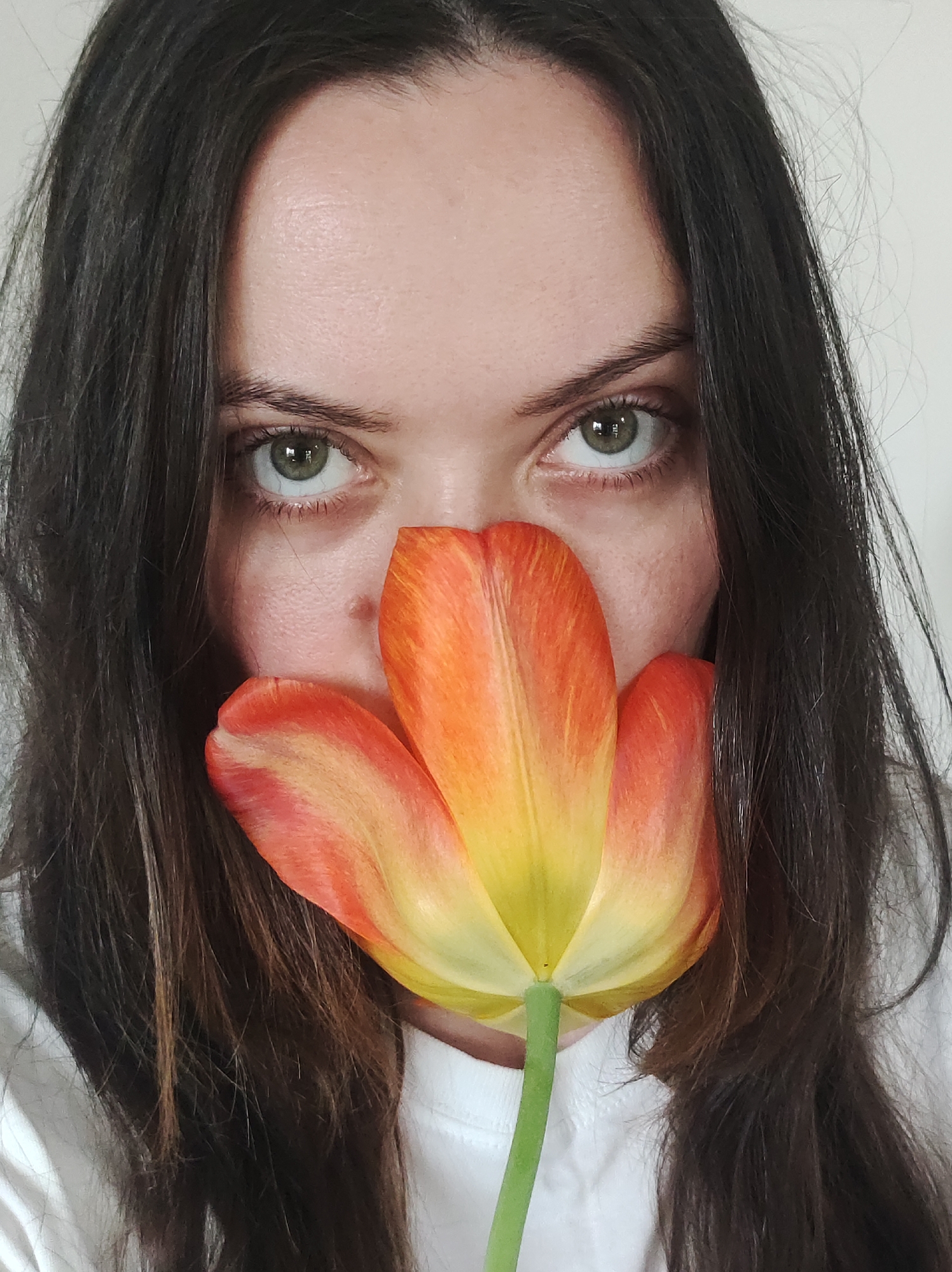 A woman with dark brown hair and green eyes is holding a tulip flower in front of her face, covering her nose and mouth. The tulip has a blend of yellow and orange hues in its petals. She is wearing a white T-shirt. The background is a plain white wall. The location is Unknown, Unknown.