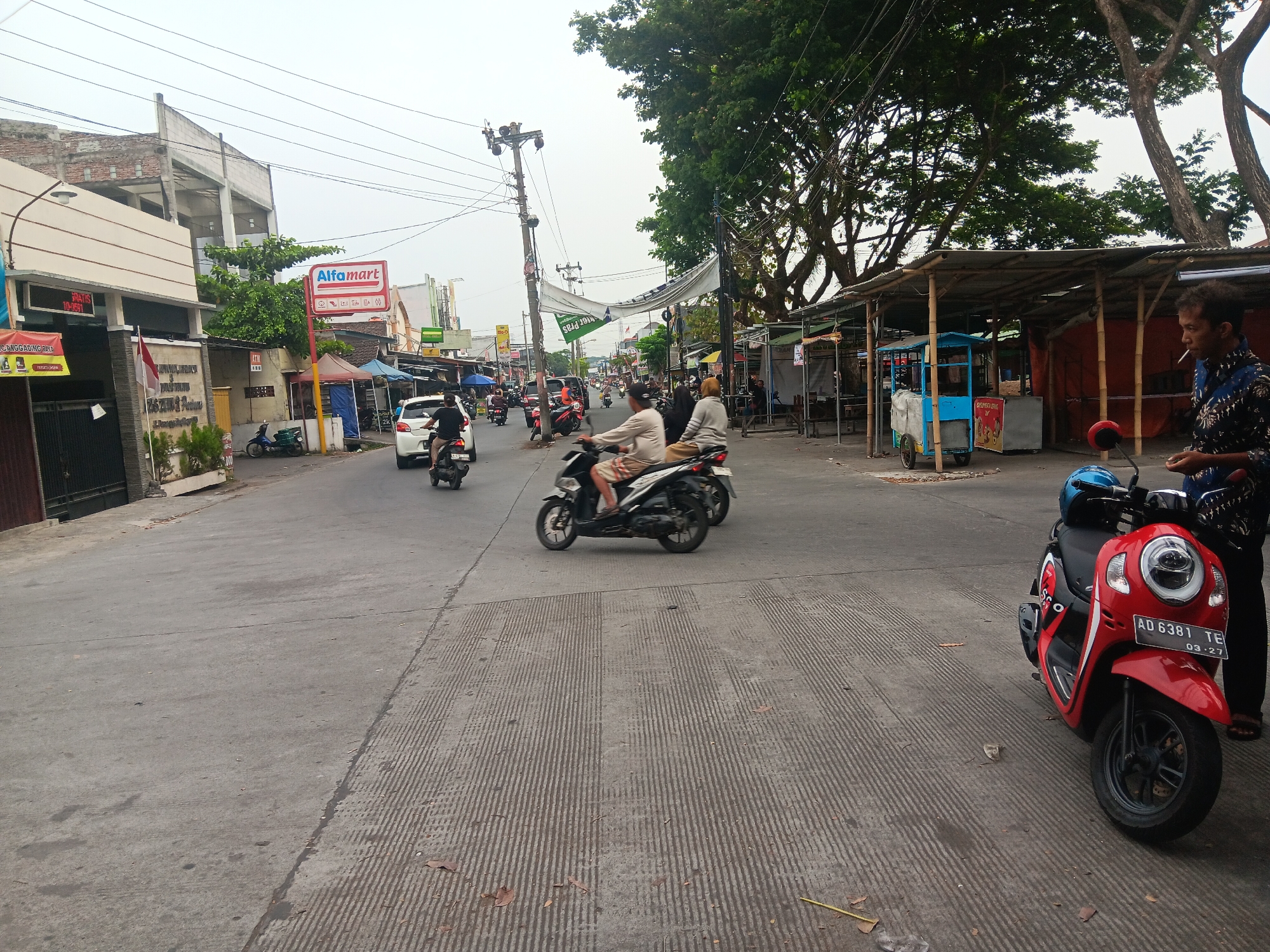 The image depicts a street scene in Batursari, Indonesia, likely during daylight hours under an overcast sky. The street is paved with asphalt, with distinct grooved sections on the right side. A variety of vehicles, predominantly motorcycles, are present, suggesting moderate traffic.
On the left side of the street, a building with an "Alfamart" sign is visible, indicating a commercial area. Adjacent to it are several shops or stalls with awnings made of corrugated metal or bamboo. These stalls appear to be selling various goods, with some displaying colorful banners and signage.
In the foreground, a red scooter with a blue helmet on the seat is parked. A man, wearing a dark patterned shirt and dark pants, is standing next to the scooter, holding a cigarette. His right hand is raised near his mouth, and he appears to be in the process of smoking.
Further down the street, several individuals are seen riding motorcycles. One motorcycle in the mid-ground has two occupants, a driver and a passenger, both blurred due to motion. Another motorcycle to the left has a single rider. In the distance, more vehicles and structures line the street, indicating a populated urban or suburban environment.
The environment is characterized by a mix of commercial buildings, roadside stalls, and trees. Power lines are visible overhead, typical of an Indonesian streetscape. The overall impression is one of a busy, everyday street scene in a localized Indonesian community.