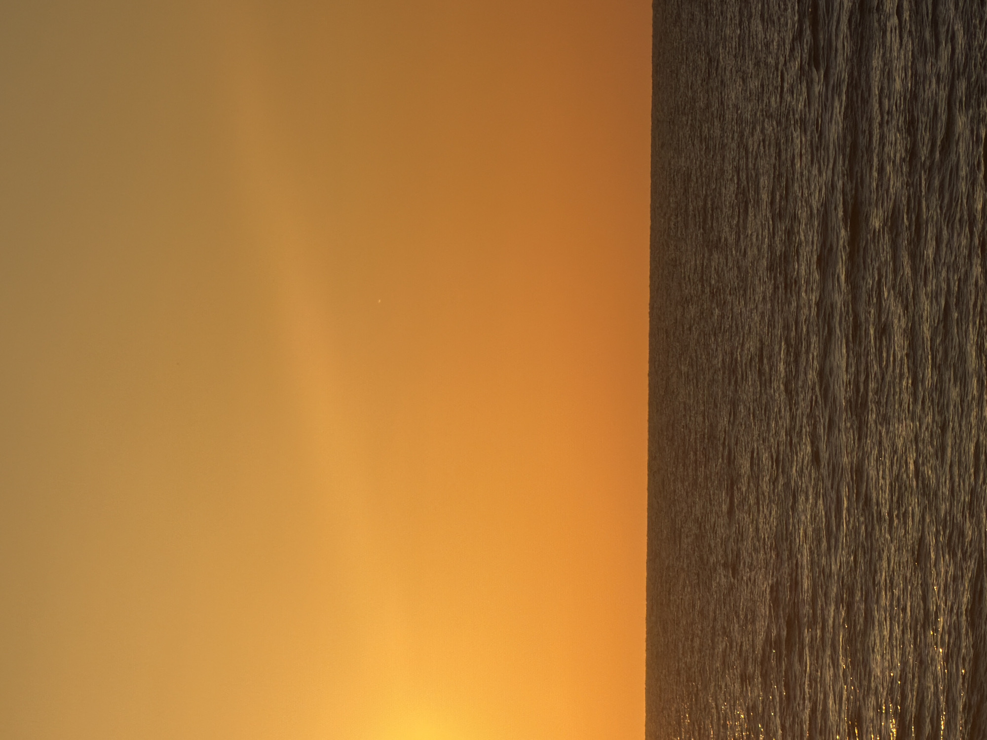 The image displays a serene landscape at what appears to be either sunrise or sunset over a large body of water. The scene is split horizontally into two distinct sections.
The upper portion of the image is dominated by the sky, painted in rich, warm hues of orange and golden yellow, characteristic of the "golden hour." The color is most intense near the horizon, gradually softening upwards. A very small, bright white dot, possibly a distant star or planet, is faintly visible in the upper left area of the sky.
The lower half of the image shows a vast expanse of water, consistent with the Mediterranean Sea given the Tel-Aviv, Israel context. The water's surface is textured with numerous small, choppy waves or ripples. These ripples catch and reflect the warm light from the sun, creating glittering streaks and patterns across the water, enhancing its dynamic appearance. The light also suggests a low sun angle, reinforcing the impression of dawn or dusk.
The overall atmosphere is tranquil and devoid of human presence or activity. There are no people, man-made objects, or discernible structures visible in the frame. The weather appears clear with no clouds. No visible text is present.