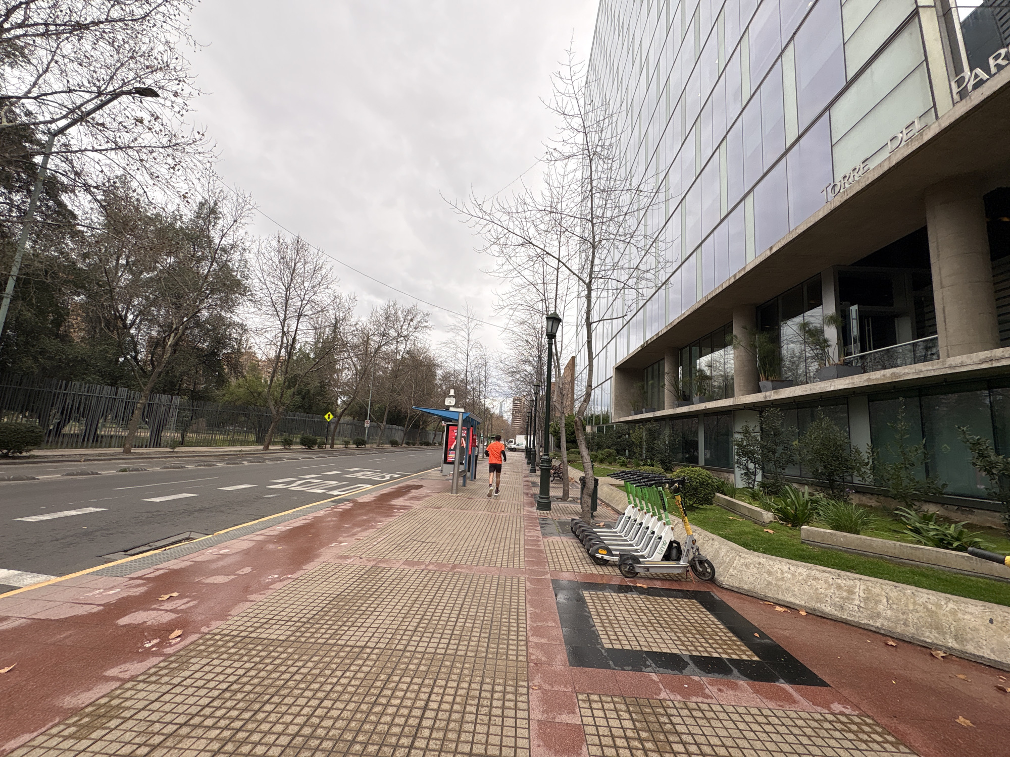 The image captures a street scene in Santiago, Chile, on an overcast day. A man in an orange t-shirt and black shorts is jogging away from the camera on a paved sidewalk. To his left is a bus stop shelter with advertisements. The sidewalk has a red and tan tile pattern, with fallen leaves scattered on its surface. A row of electric scooters is parked on the sidewalk to the right of the jogger. Beyond the sidewalk, a street with white lane markings and a yellow curb is visible, with a traffic barrier on the far side. A modern glass-fronted building, identified by signage as "TORRE DEL PARQUE," dominates the background to the right. Trees with bare branches line the street and sidewalk, suggesting a cooler season. The sky is cloudy and light.