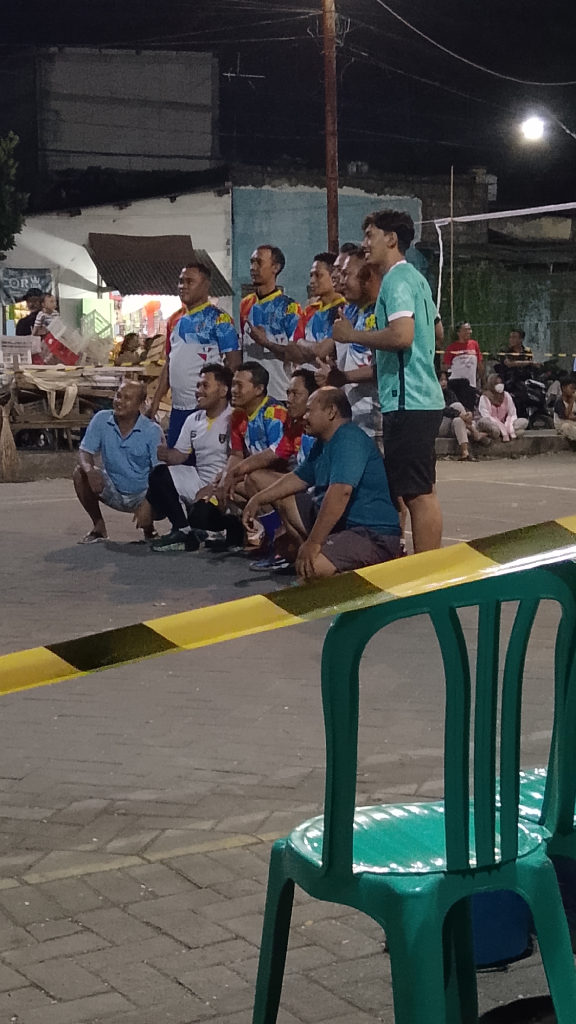 In Banjarsari, Indonesia, a group of men, some posing for a photo, are gathered on a paved outdoor area at night. The group consists of at least ten individuals, mostly young to middle-aged men, wearing athletic attire, including jerseys with colorful designs. Several men are squatting in the front row, while others stand behind them. One man on the far right, wearing a teal jersey, is giving a thumbs-up. In the background, there are buildings, a volleyball net, and other people observing the scene. Two green plastic chairs are positioned in the foreground, with a yellow and black striped caution tape barrier in front of them. The lighting suggests it is nighttime, possibly illuminated by artificial lights.
