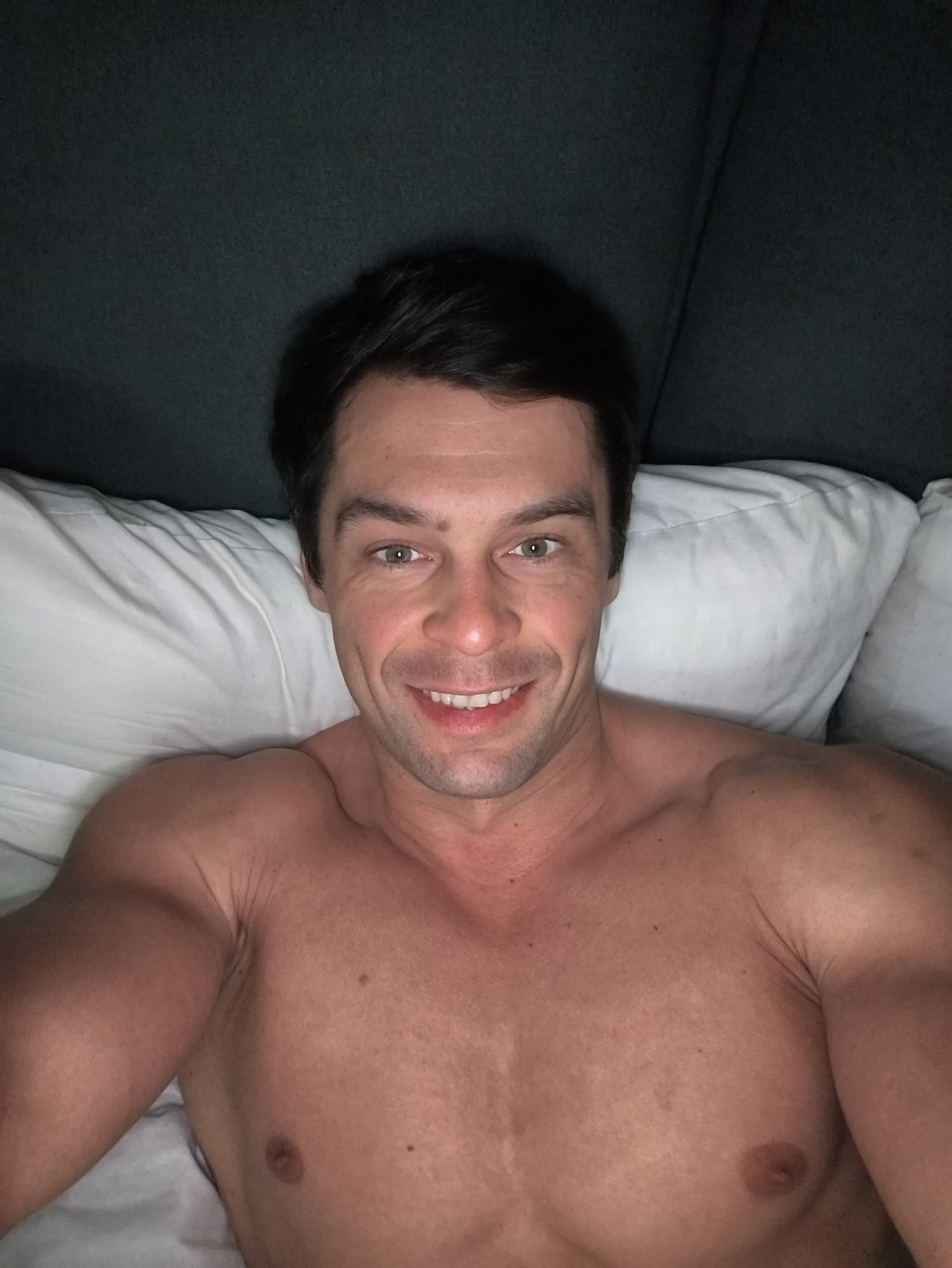 A shirtless man is lying in bed, smiling at the camera. He is positioned between white pillows, and behind him is a dark headboard. The location is not discernible from the image.