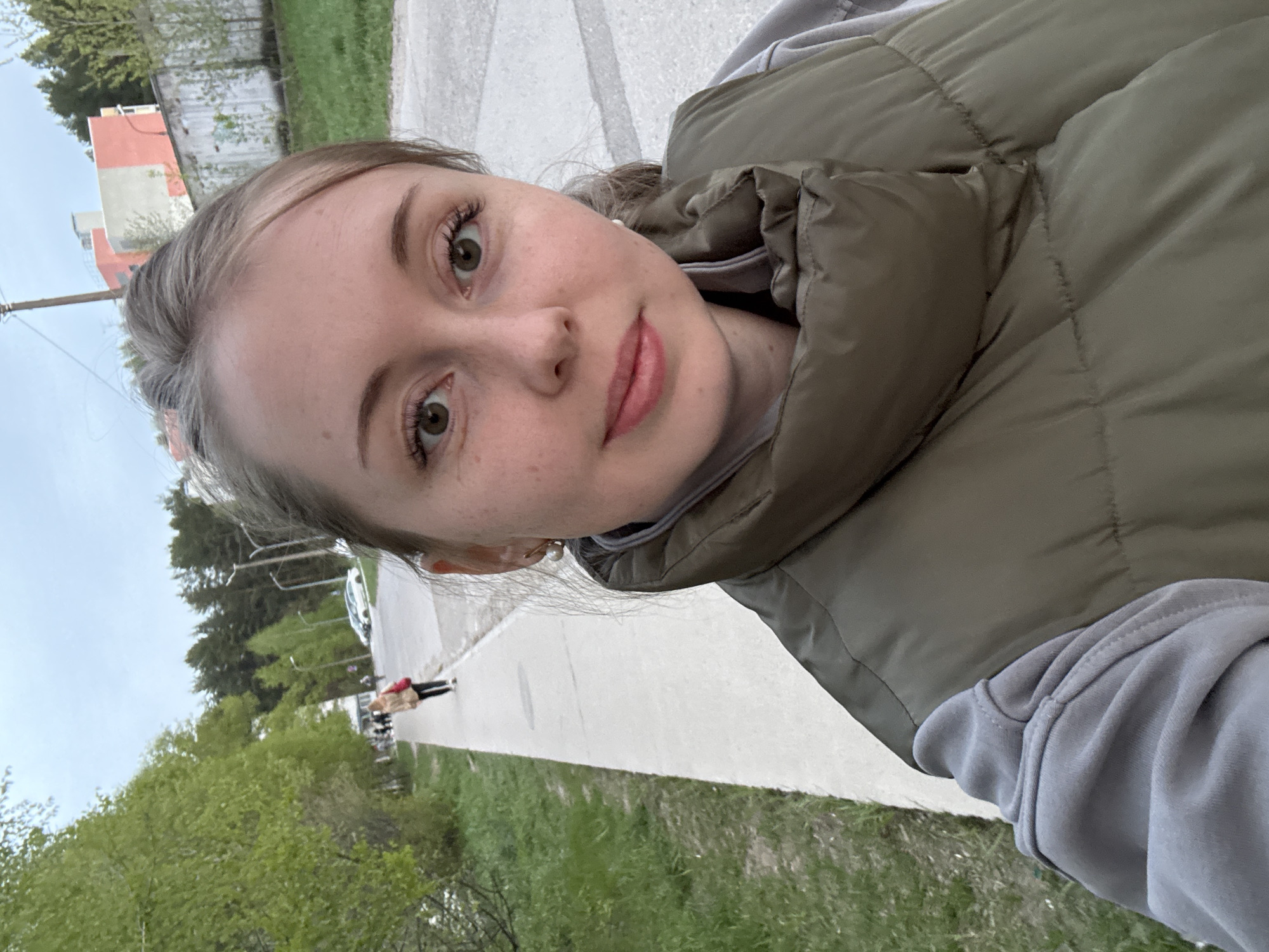A close-up selfie of a young woman wearing a gray hooded sweatshirt and an olive green puffy vest. She has light skin, freckles, and light brown hair pulled back. Behind her is a sidewalk, a grassy area, and a street with buildings and trees in the distance. There are other people walking down the sidewalk. The location is the city of Unknown, Unknown.