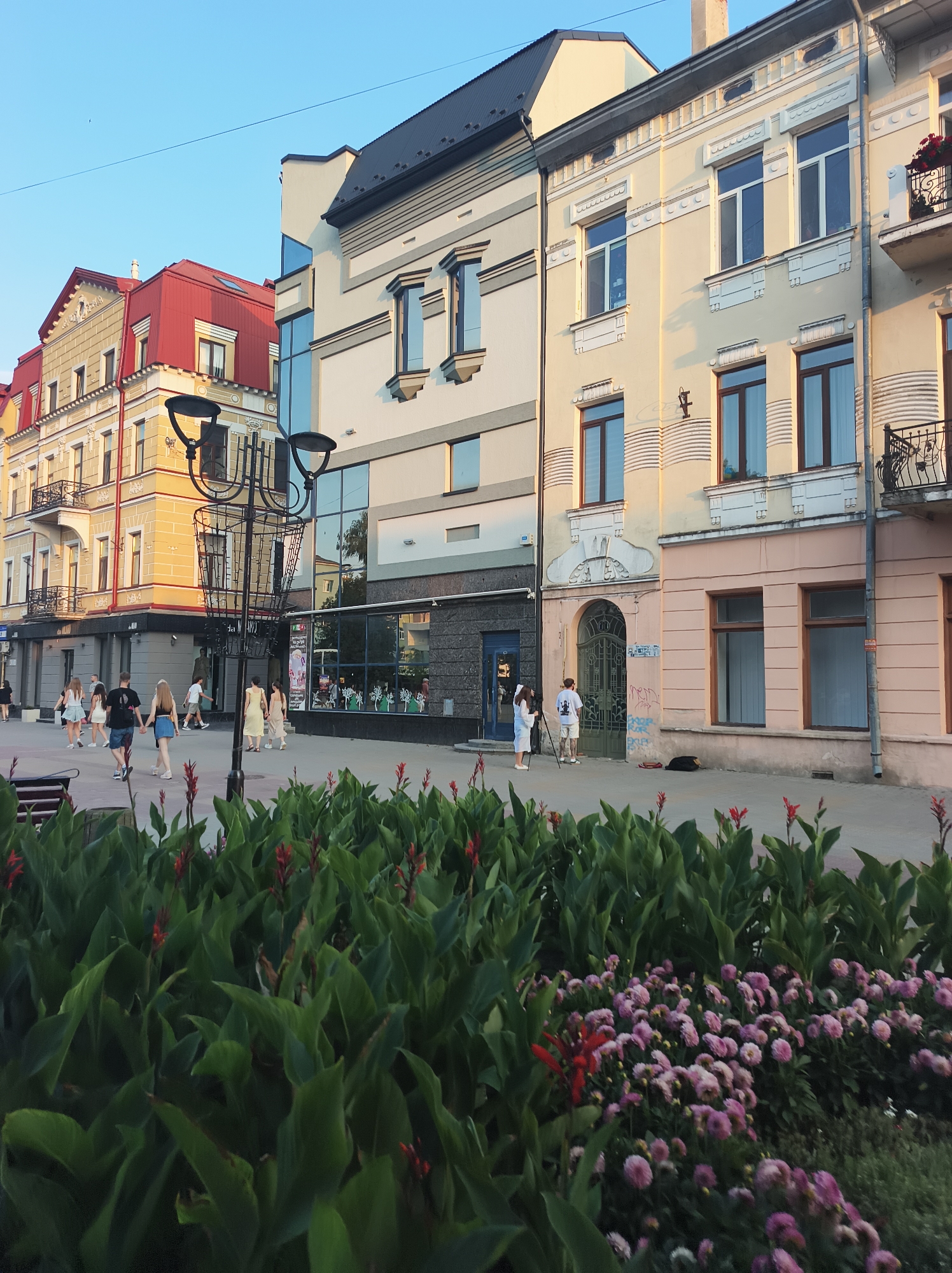 The image captures a street scene in Ternopil, Ukraine, likely during a late afternoon or early evening. In the foreground, lush green foliage and pink flowers create a vibrant border. Beyond the greenery, a paved walkway is visible with several people strolling. On the left, a group of young people, two couples and another individual, are walking together. Further down the walkway, a person in a yellow dress walks away from the camera, followed by another person in a light-colored dress. A tall, ornate lamppost stands prominently in the middle ground.
Behind the walkway, a row of multi-story buildings lines the street. The architecture varies, with the leftmost building featuring a striking red roof and ornate yellow facade. Adjacent to it is a modern building with a grey and white exterior and large glass windows. The building to the right of that is painted a pale yellow with white decorative elements around the windows and an arched entryway. The building furthest to the right has a lighter pink facade.
The sky above is a clear, light blue, indicating good weather. The overall impression is one of a pleasant urban environment with people enjoying a leisurely walk on a well-maintained street. The scene suggests a typical day in the city.