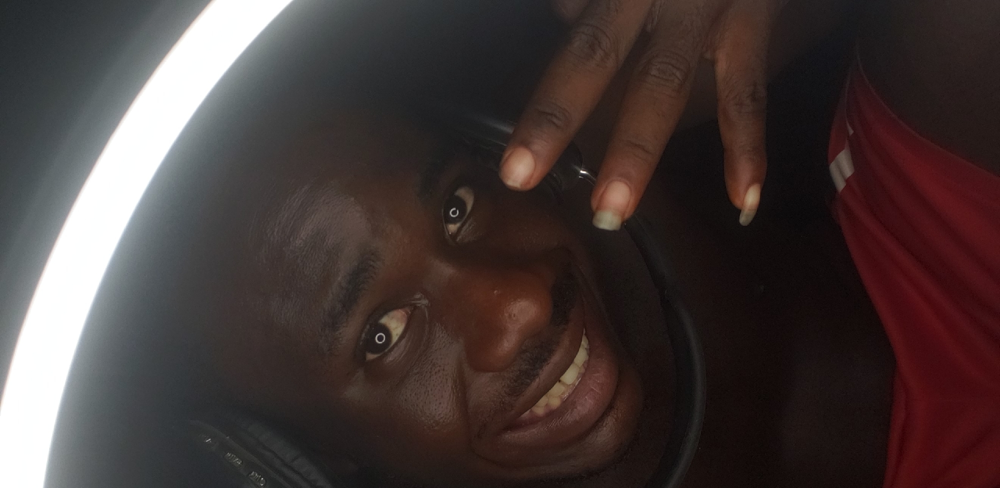 A close-up, angled portrait of a dark-skinned person with a shaved head, located in Narayi, Nigeria. The person is smiling, revealing teeth, and wearing headphones. A hand with long nails is positioned near the face. The background is dark, with a circular white light source partially visible on the left. A red garment is seen in the lower right corner.