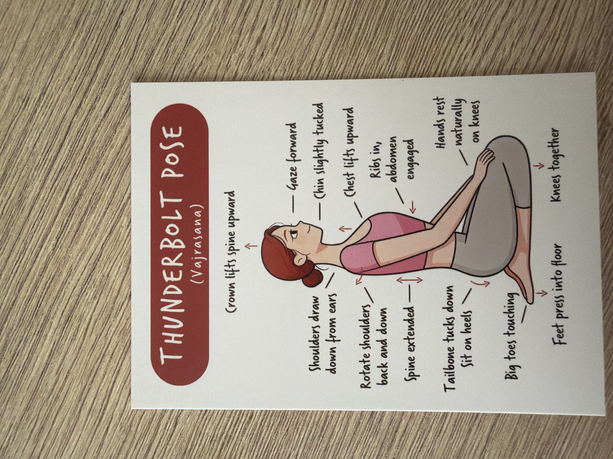 The image is a close-up, eye-level photograph of a yoga pose instruction card. The card is white with red and black text and a cartoon illustration of a woman in the Thunderbolt Pose (Vajrasana). The card is placed on a light brown wooden surface with visible grain. The title "THUNDERBOLT POSE (Vajrasana)" is in red at the top of the card. To the right of the illustrated woman, there are several bullet points describing the pose: "Gaze forward," "Chin slightly tucked," "Chest lifts upward," "Ribs in, abdomen engaged," and "Hands rest naturally on knees." To the left of the woman, there are more instructions: "Crown lifts spine upward," "Shoulders draw down from ears," "Rotate shoulders back and down," "Spine extended," "Tailbone tucks down Sit on heels," "Big toes touching," and "Feet press into floor." The illustration depicts the woman with her legs folded underneath her, sitting on her heels. Her back is straight, and her head is slightly tilted. Her arms are resting on her knees, and her feet are shown with the big toes touching. The lighting is even, and the focus is sharp on the card. There is no information or visual element within the image that relates to the city of Pazardzhik, Bulgaria.
