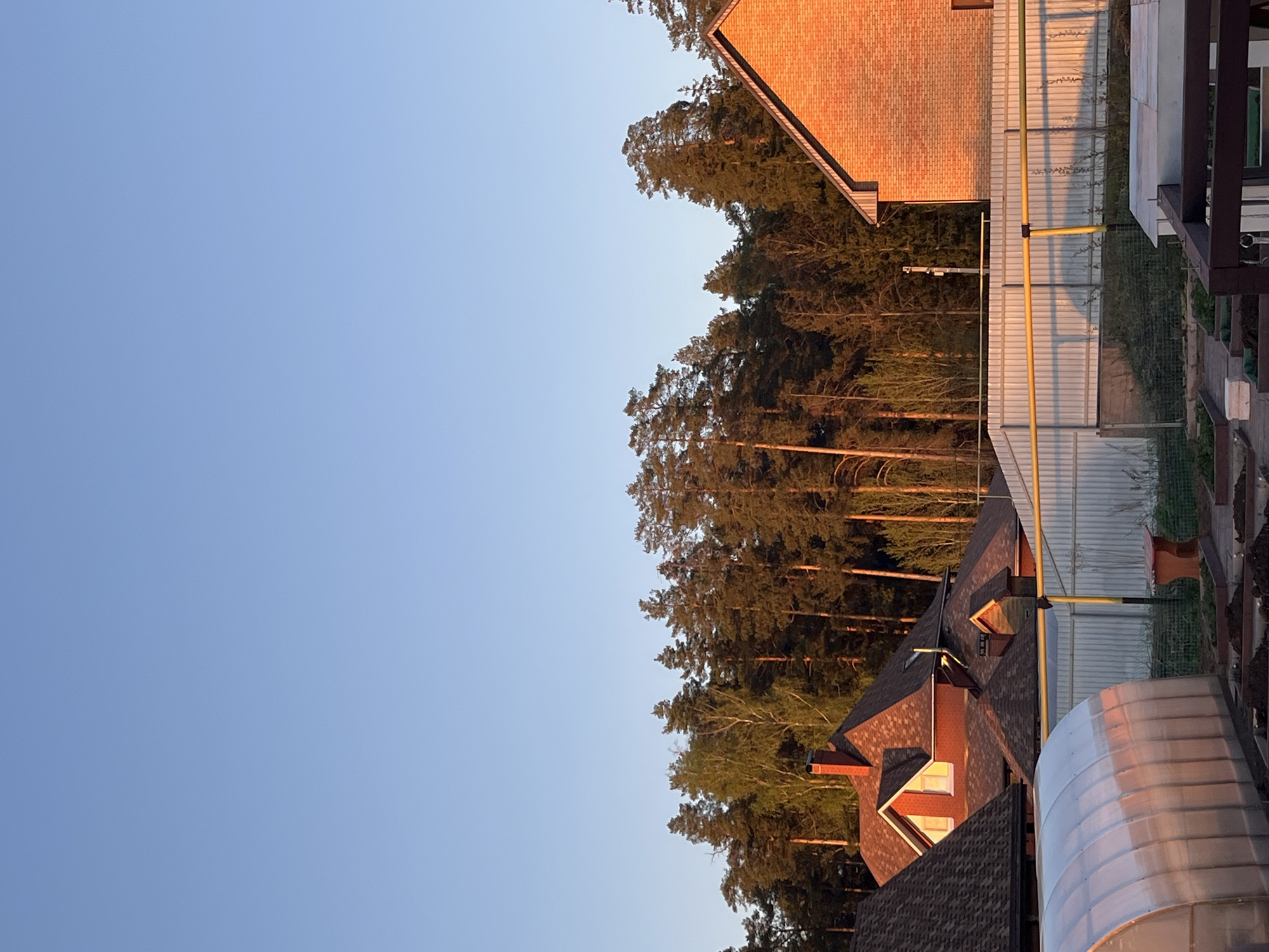 The image shows a residential area in Chaikovsky, Russia, taken at dusk or dawn. The sky is a gradient of light blue. Dense trees line the horizon. Several buildings are visible. A brick house with a gable roof stands adjacent to a lighter-colored house. Corrugated metal fencing separates the houses and runs along the edge of the property. A yellow pipe stands next to the fence. A silver, corrugated metal structure of an unknown use sits in the foreground.