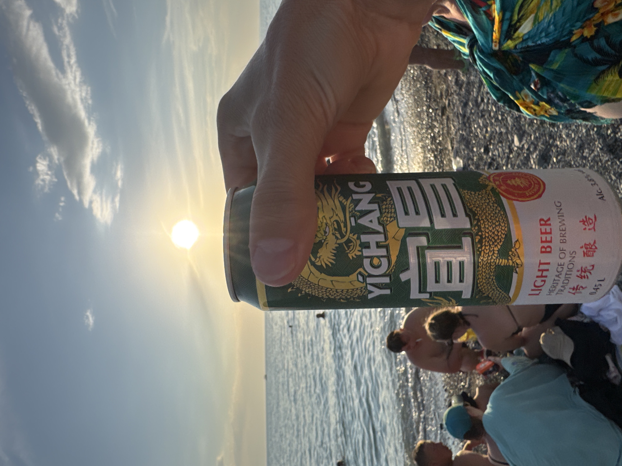 A first-person perspective shot shows a hand holding a Yichang Light Beer can against a beach scene at sunset. The sun is visible in the sky, casting a warm glow. The ocean is in the background, with several people visible in the water and on the shore. The can itself is green with a dragon design and text in English and Chinese. The beach appears to be covered in pebbles. The presence of a beach and ocean suggests a coastal or lakeside location. The image does not provide information about the city of Zharbulak, Kazakhstan, or any details that would place it there specifically.