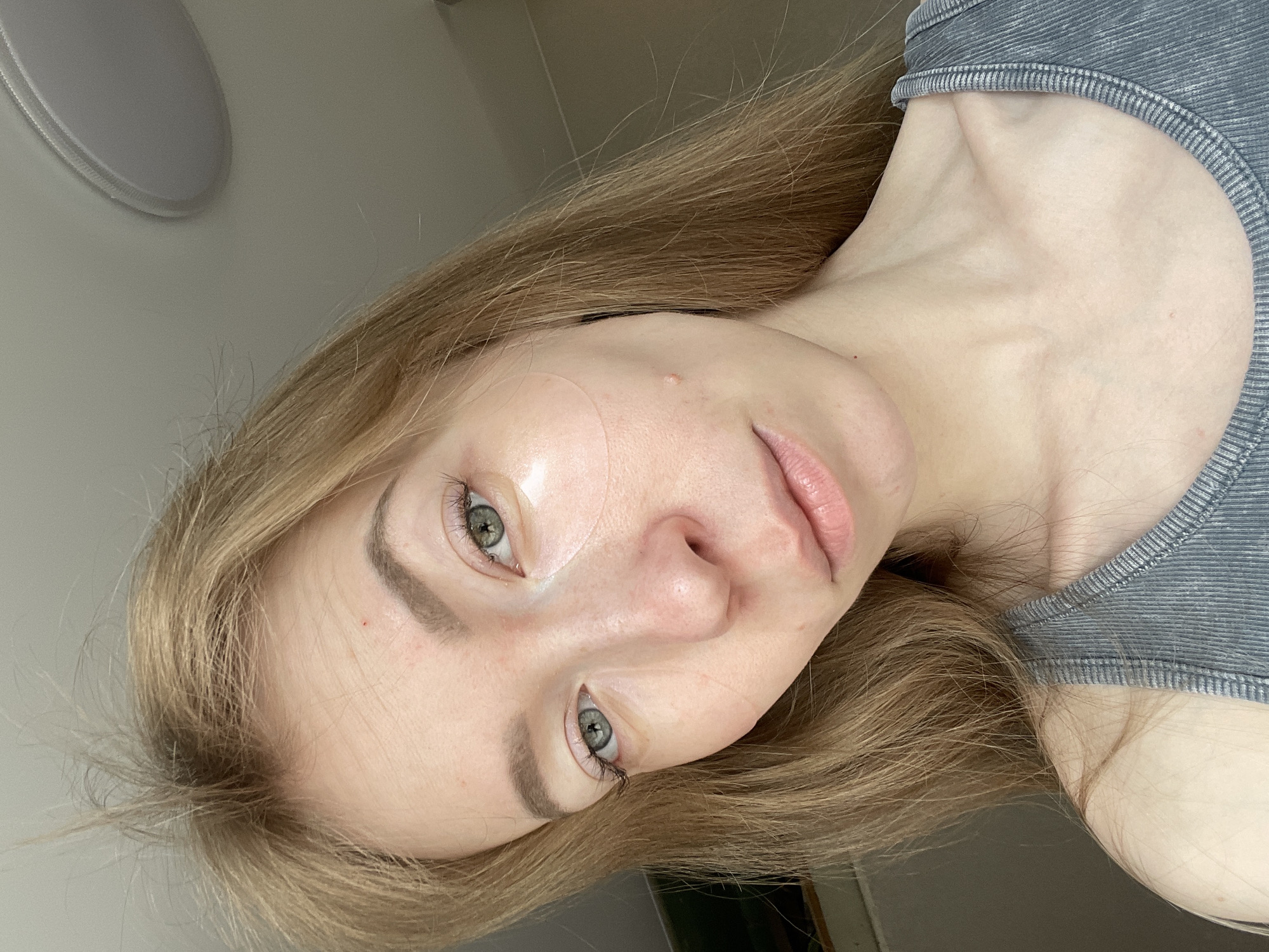 The image shows a fair-skinned woman with blonde hair. She is wearing a gray tank top. There are translucent facial patches on her forehead. The background includes a plain white wall with a circular light fixture. The location, Perm, Russia, is not discernible from the image.