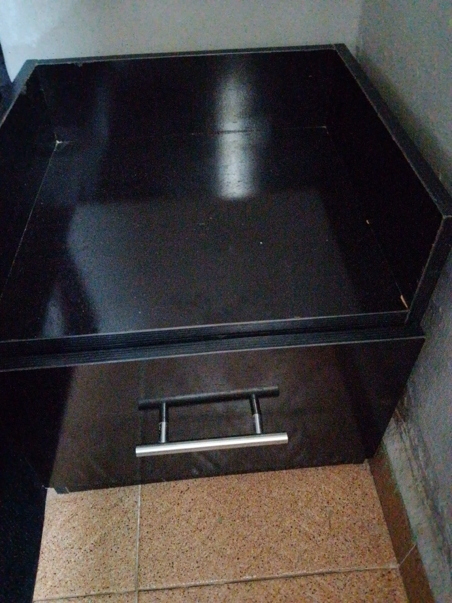 A top-down, close-up shot shows a dark, rectangular object with a drawer. The drawer has a silver handle. The object appears to be made of wood or laminate. The background is out of focus, but it appears to be a wall and floor.