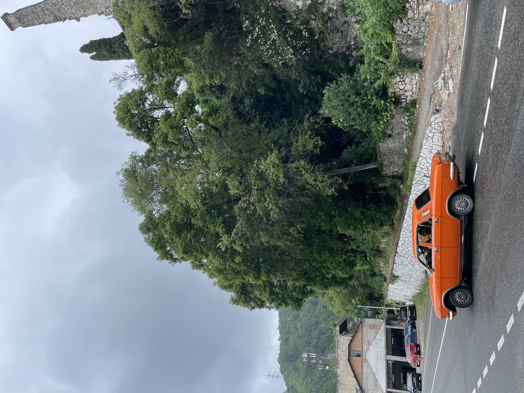 An exterior, medium shot captures a bright orange sedan driving on a wet asphalt road under an overcast sky in Abaata, Abkhazia. The car is positioned in the right foreground, facing right, with its driver visible inside. Behind the car, a stone wall with a decorative pattern runs along the side of the road, separating it from a steep, tree-covered embankment. A light-colored building, possibly a workshop or garage, is situated on the left side of the road, with several other vehicles parked in front of it. The background is dominated by lush green trees and a distant, hazy hillside.