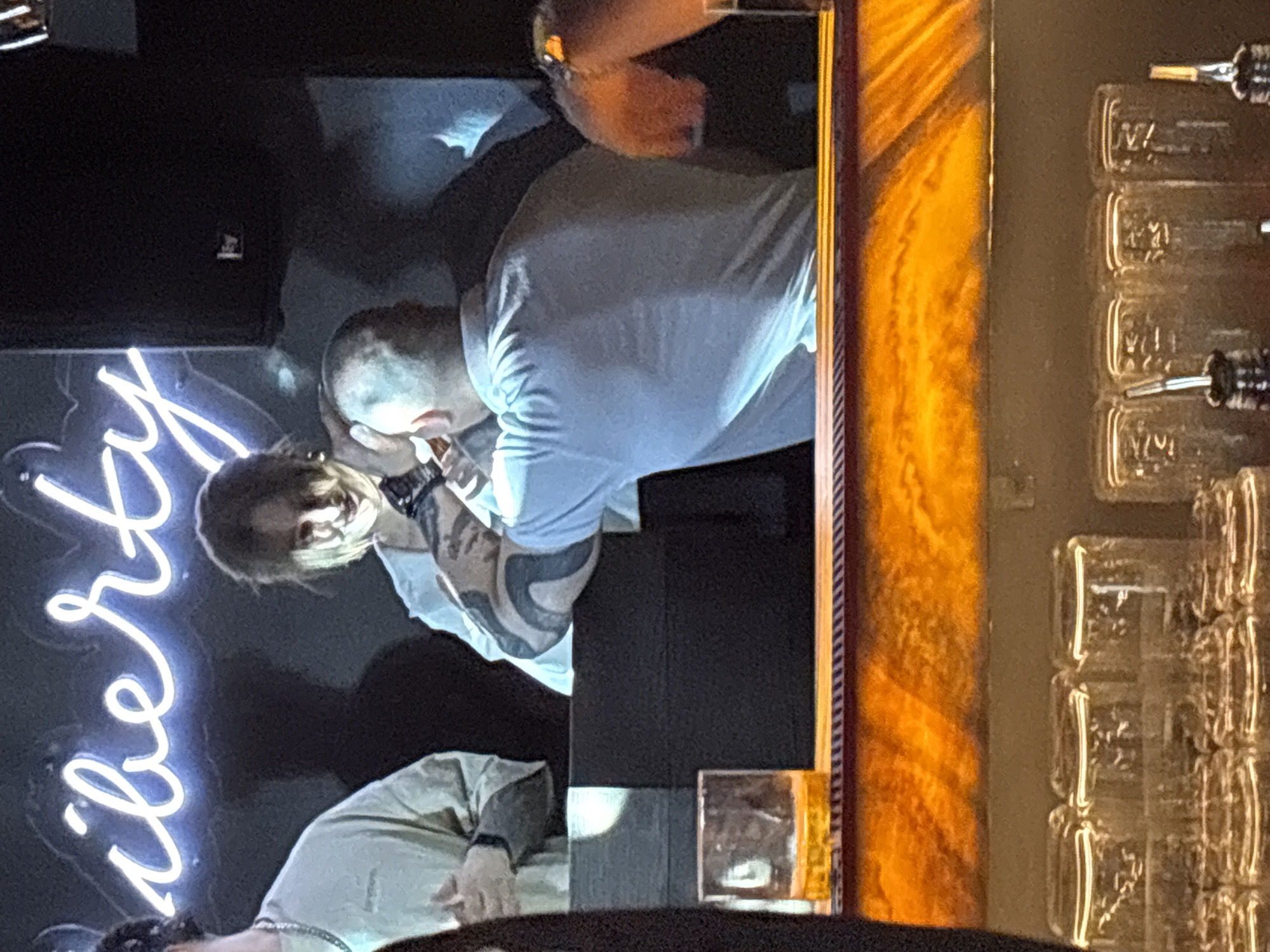 A scene from Liberty Bar in Perm, Russia: a person with tattoos is seated at a bar, next to a bald person in a white shirt. There is a neon sign that says “Liberty” in the background, and a row of glassware next to them on the bar.