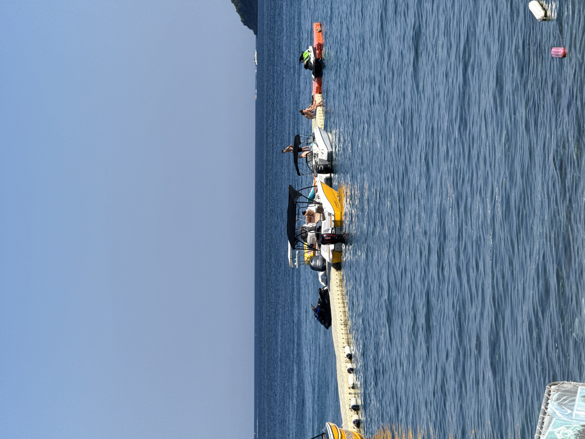 The image depicts a sunny day scene in Budva, Montenegro, with a focus on a floating pontoon dock extending into the calm, blue sea. Several boats and jet skis are moored to the dock. In the foreground, a yellow boat is partially visible on the left. Further along the dock, a white boat with a black canopy is anchored, with people visible aboard and around it. To the right of this boat, a smaller orange floating platform is visible, with two individuals sitting on it. A green and white jet ski is also attached to the orange platform. In the distance, the Montenegrin coastline is visible, with a wooded hill sloping down to the sea under a clear blue sky. Several other boats are scattered across the horizon. A buoy and a smaller pink floating object are present in the water to the right of the image.