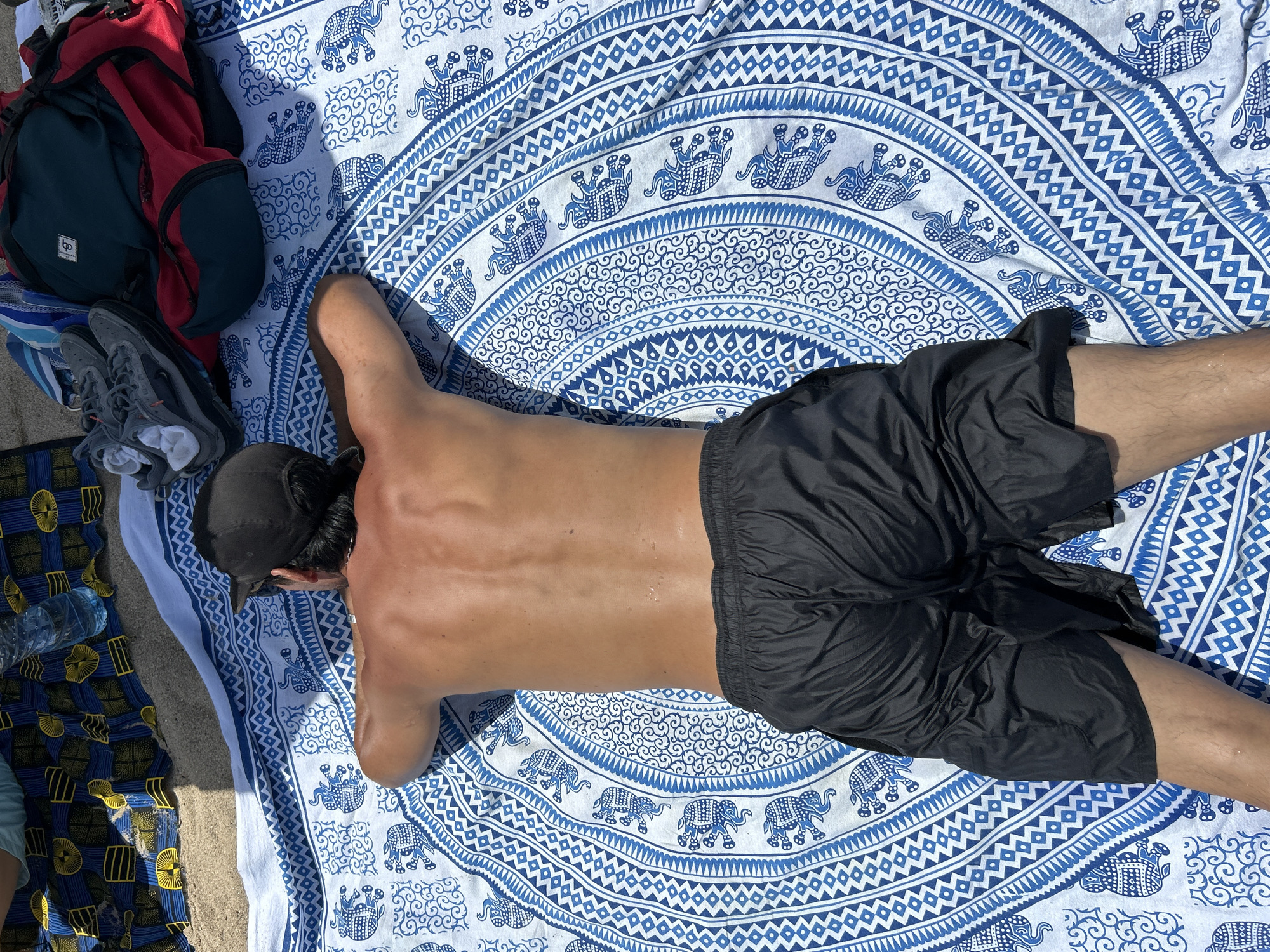 A person is lying face down on a blue and white mandala-patterned beach blanket, on a sandy surface. The person is wearing black shorts and a black baseball cap. Their back is exposed and appears tanned. To the upper right of the person, there is a red and green backpack and a pair of grey sneakers with white socks. Further to the left, there is a blue and yellow patterned cloth and a plastic water bottle. The scene is brightly lit by sunlight. The setting appears to be a beach, possibly in Barcelona, Spain.